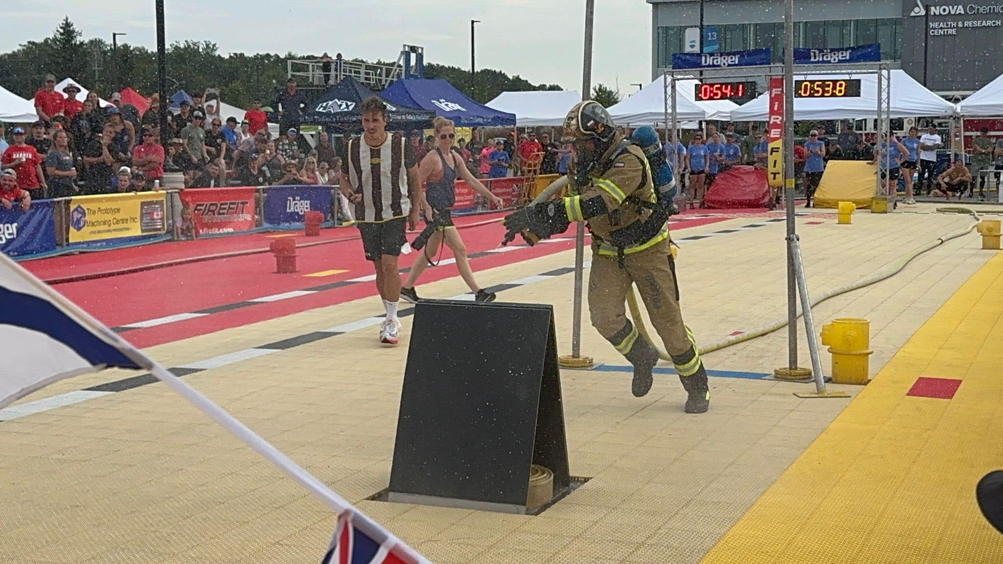 Zawodnik w mundurze strażackim biegnie podczas zawodów , obok niego mężczyzna i kobieta w strojach sportowych, w oddali tablica sportowa z wyświetlonymi wynikami 0;54.1 oraz 0;53.8