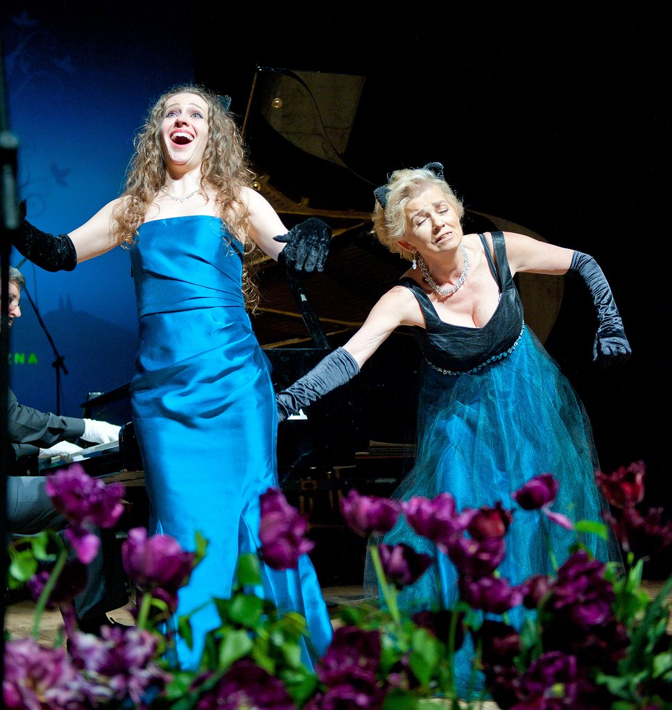 Zdjęcie. Jubileusz 60-lecia PSM w Nysie - II Koncert Jubileuszowy - 14 maja 2022 - Nyski Dom Kultury. Na scenie w trakcie śpiewania od lewej Katarzyna Santi Sperczyńska i Jadwiga Chlewicka. W tle z lewej strony przy fortepianie Janusz Dąbrowski. Z przodu rozmyte bordowe kwiaty. Kliknięcie na zdjęcie przeniesie do prezentacji cyklu zdjęć z koncertu.