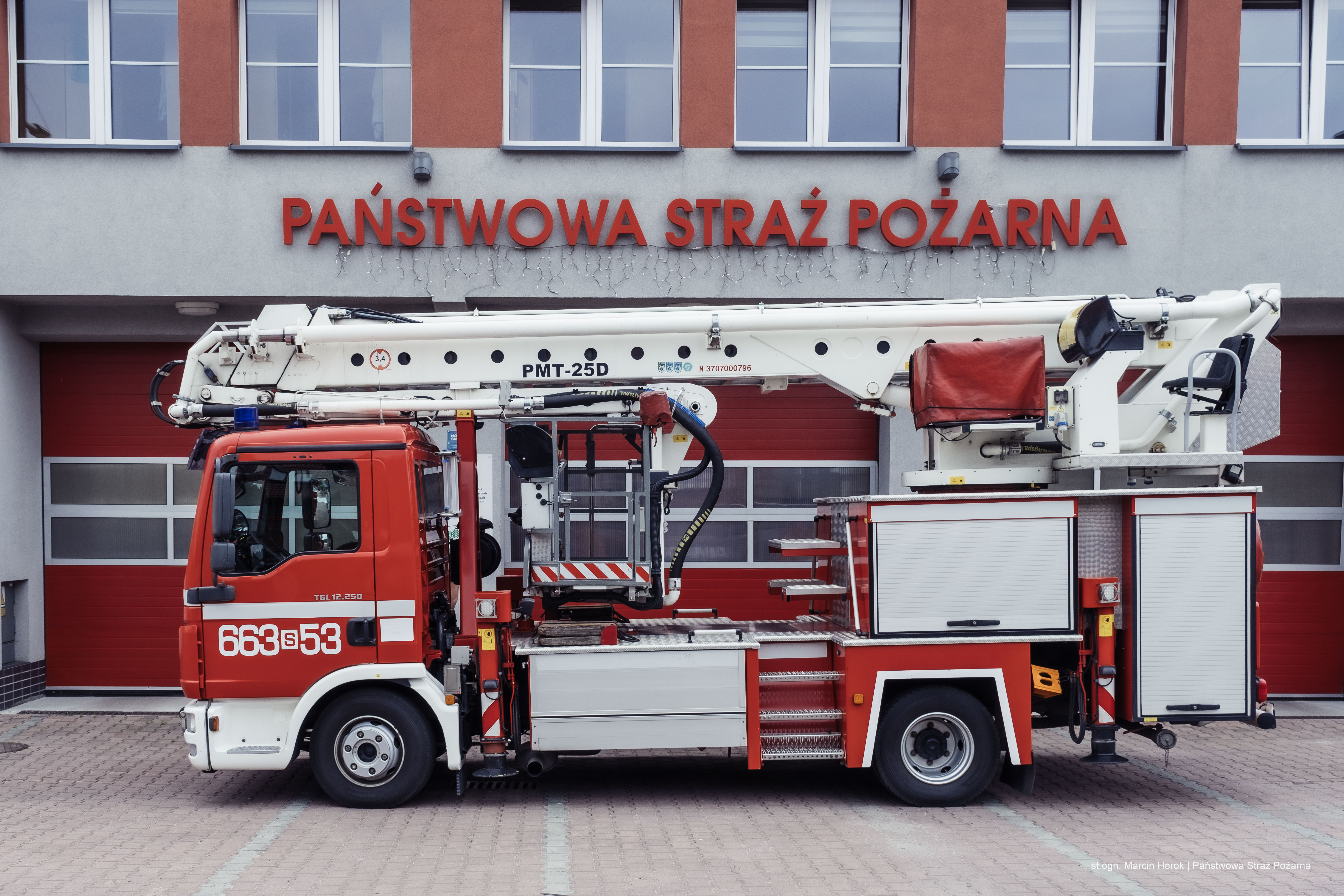 Zdjęcie przedstawia samochód specjalny z podnośnikiem hydraulicznym o wysięgu 25 metrów SH 25D. W tle budynek Jednostki Ratowniczo-Gaśniczej w Rydułtowach.