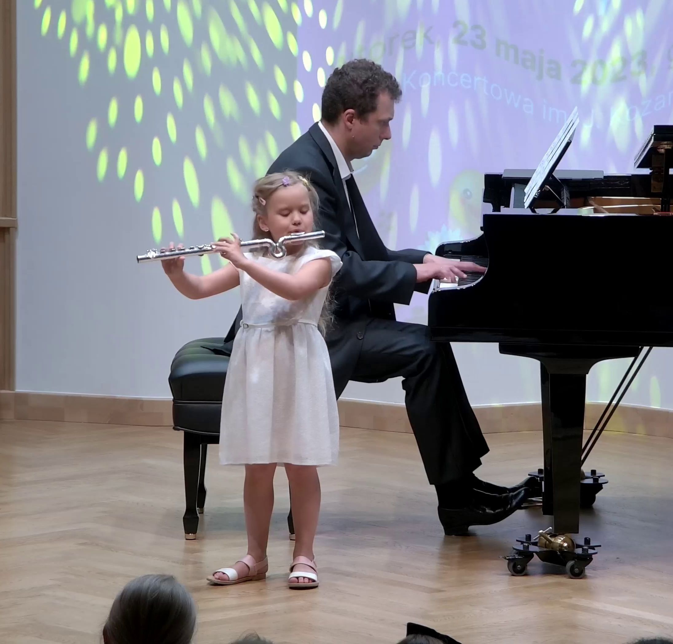Zdjęcie. Na estradzie Sali Koncertowej występuje mała flecistka. Towarzyszy jej przy fortepianie dorosły pianista, grający na dużym czarnym fortepianie.