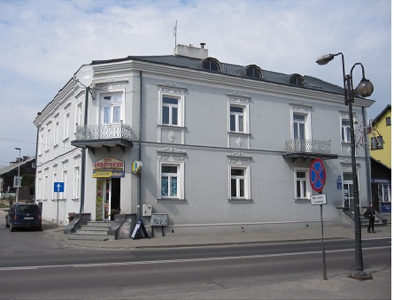 Zdjęcie przedstawia jasno szary narożny budynek z grafitowym dachem. Do budynku prowadzą narożne schody. 