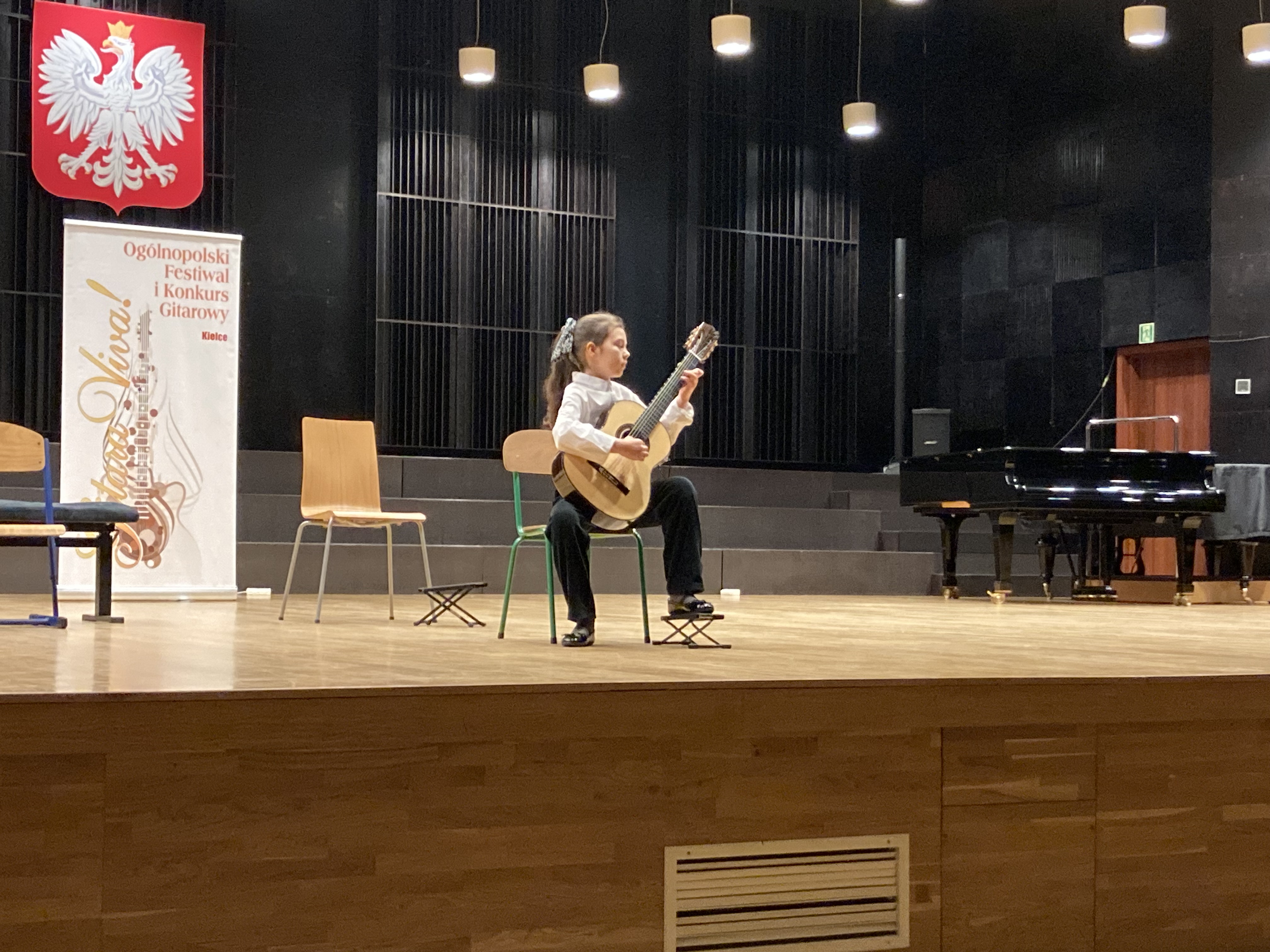 Sala koncertowa Zespołu Szkół Muzycznych w Kielcach, ogromna scena. Na środku sceny występuje uczennica naszej szkoły Lena Torba podczas X Ogólnopolskiego Festiwalu i Konkursu "Gitara Viva" w dniu 16 marca 2023 r. Po lewej stronie na ścianie wisi duże godło Polski pod nim stoi baner z napisem "Gitara Viva!" na suficie wisi mnóstwo żyrandoli. Uczennica siedzi na krześle po środku sceny, podczas występu, po jej lewej stronie stoją dwa krzesła po prawej fortepian.