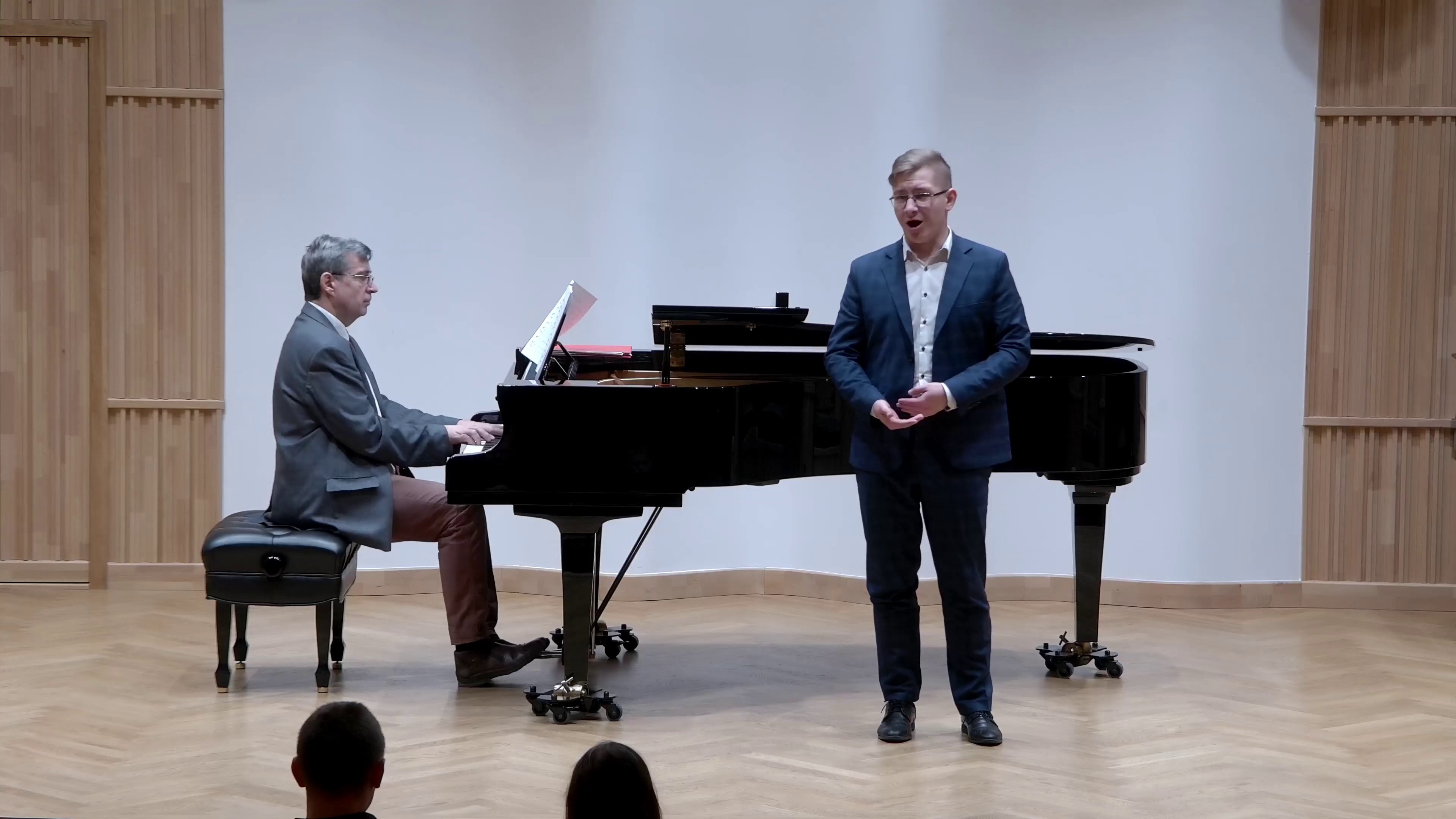 Zdjęcie. Na estradzie występuje śpiewak z towarzyszeniem pianisty, który gra na dużym czarnym fortepianie.