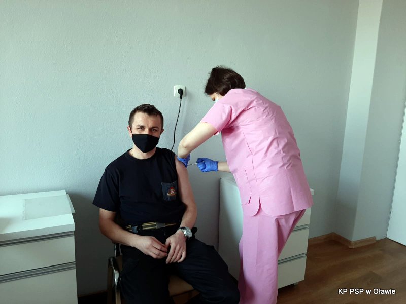 Na zdjęciu widać siedzącego strażaka w ciemnym mundurze koszarowym, który właśnie przyjmuje szczepionkę przeciw COVID-19. Po prawej stronie stoi pielęgniarka, która wykonuje zastrzyk.