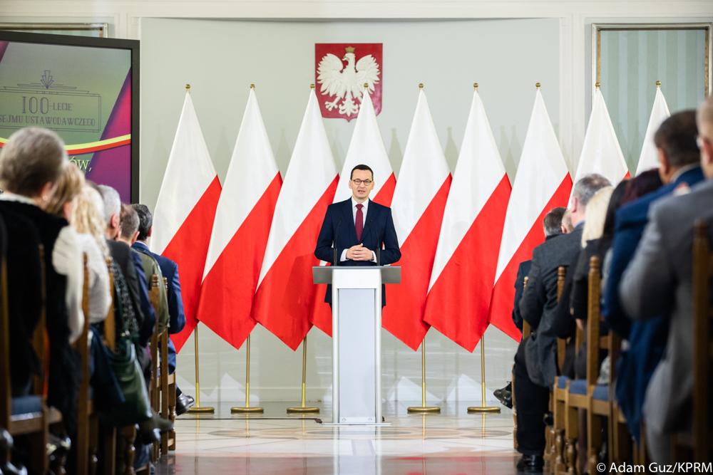 Premier Mateusz Morawiecki przemawia na tle flag Polski, a po bokach siedzą uczestnicy uroczystości.