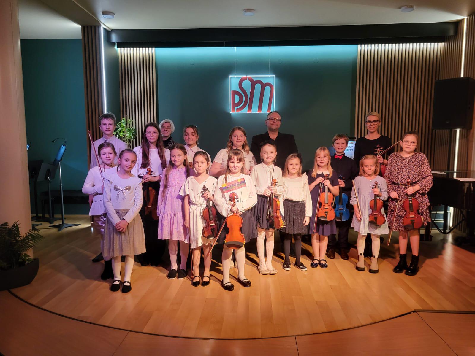 Na zdjęciu widać uczniów klas skrzypiec którzy trzymają w rękach skrzypce i klasy fortepianu. Uczniowie stoją w rzędzie a za nimi nauczyciele od skrzypiec i fortepianu poprawi stronie widoczny fragment fortepianu. W tle widoczne ściana w kolorze zielonym oraz logo szkoły PSM.
