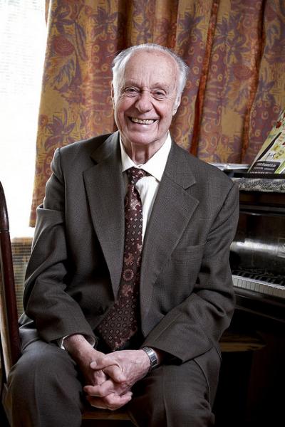 zdjęcie przedstawia w części głównej sylwetkę Patrona PSM I st. im. Andrzeja Cwojdzińskiego w Tomaszowie Lubelskim, ubranego w marynarkę, z uśmiechniętą twarzą, od góry w tle kolorowa zasłonka z elementami roślinnymi, z prawej strony kawałek części fortepianu z otwartą klawiaturą