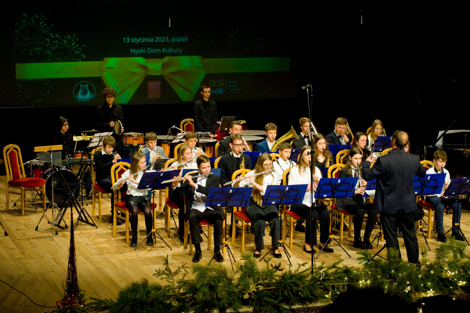 Zdjęcie. Na estradzie Sali Widowiskowej Nyskiego Domu Kultury występuje zespół instrumentów dętych, w tyle sekcja instrumentów perkusyjnych. Z przodu estrada przyozdobiona gałązkami iglaków, w tle baner promujący Koncert Noworoczny. Zespół prowadzi dyrygent.