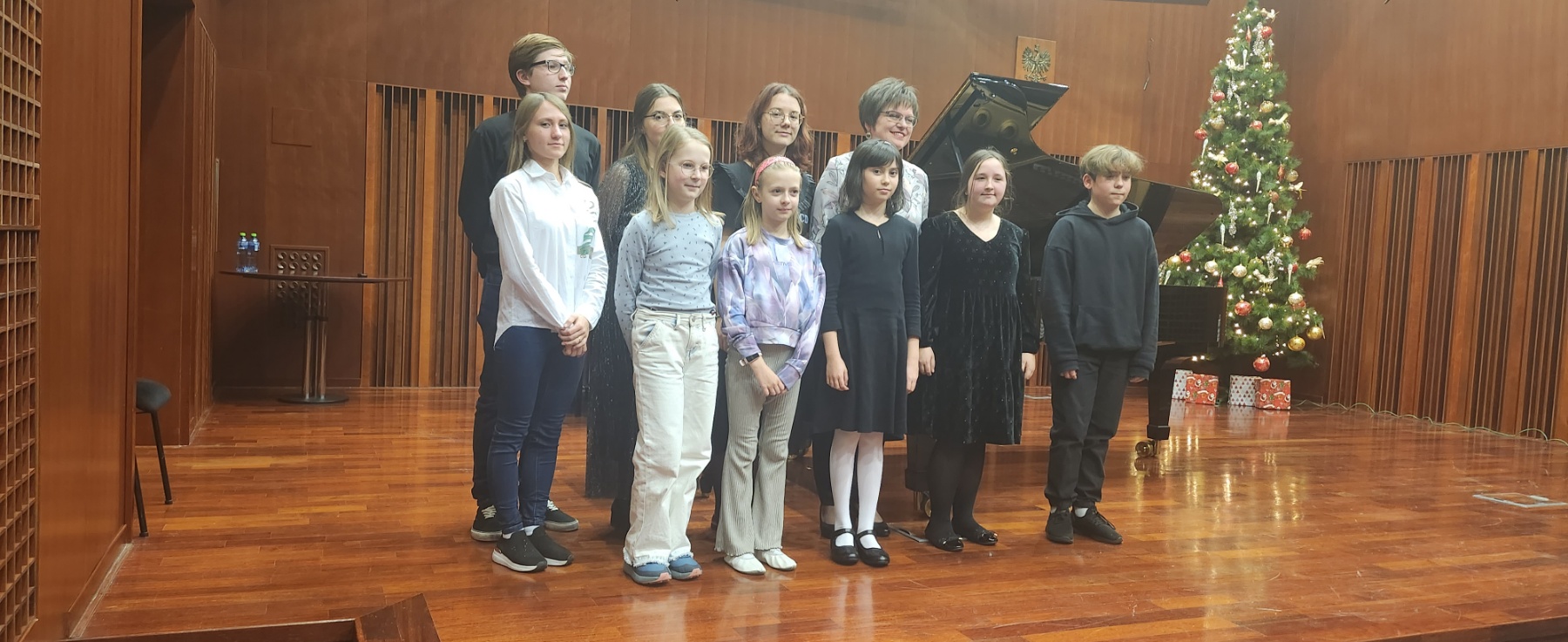 Zdjęcie uczestników warsztatów pianistycznych wraz z prof. Oksaną Rapitą w sali koncertowej szkoły. W tle brązowa scena, fortepian oraz choinka.
