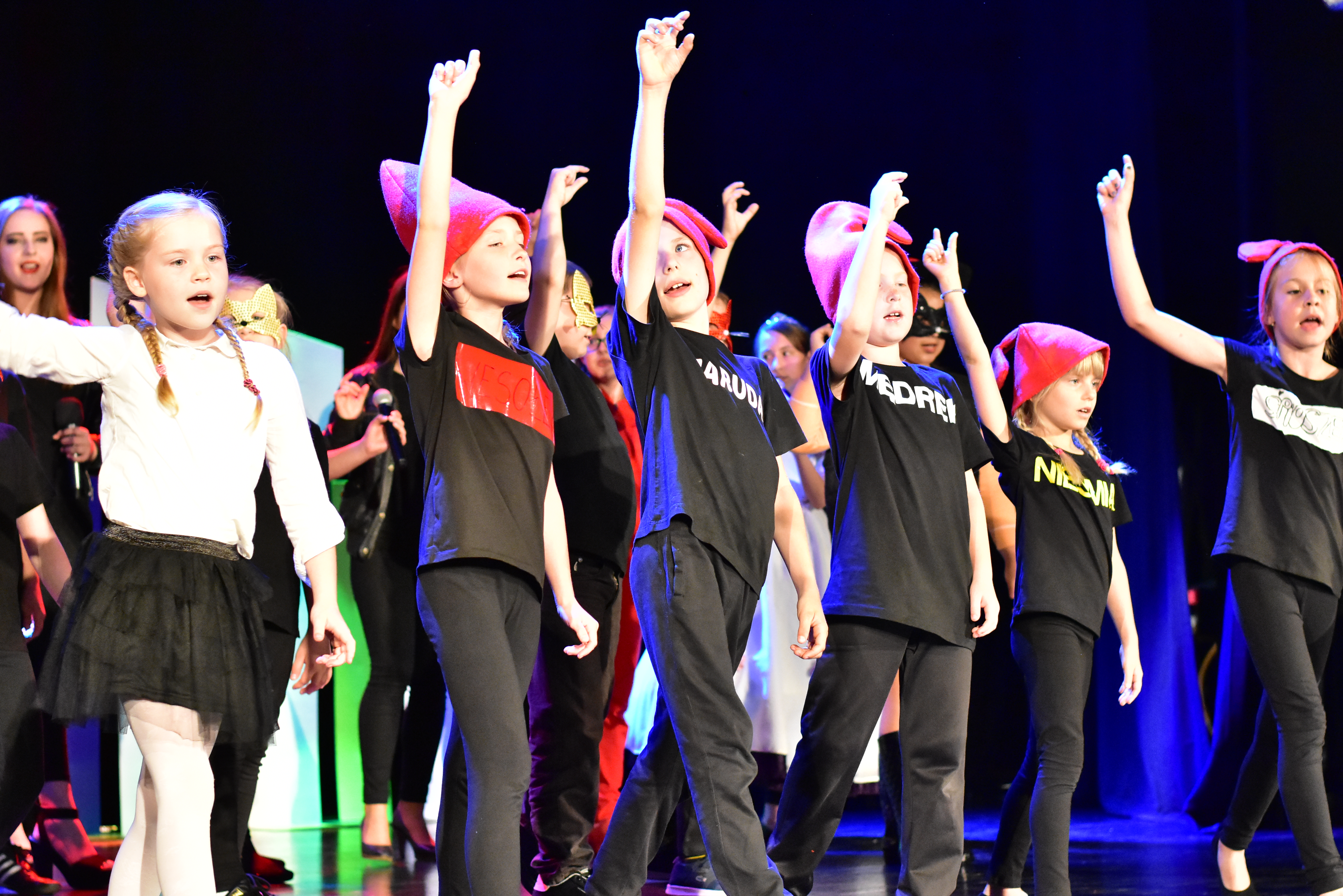 Fotografia przedstawia tańczącą grupę dzieci. Mają one czarne stroje oraz czerwone czapki. Każde z nich ma uniesioną prawą rękę.