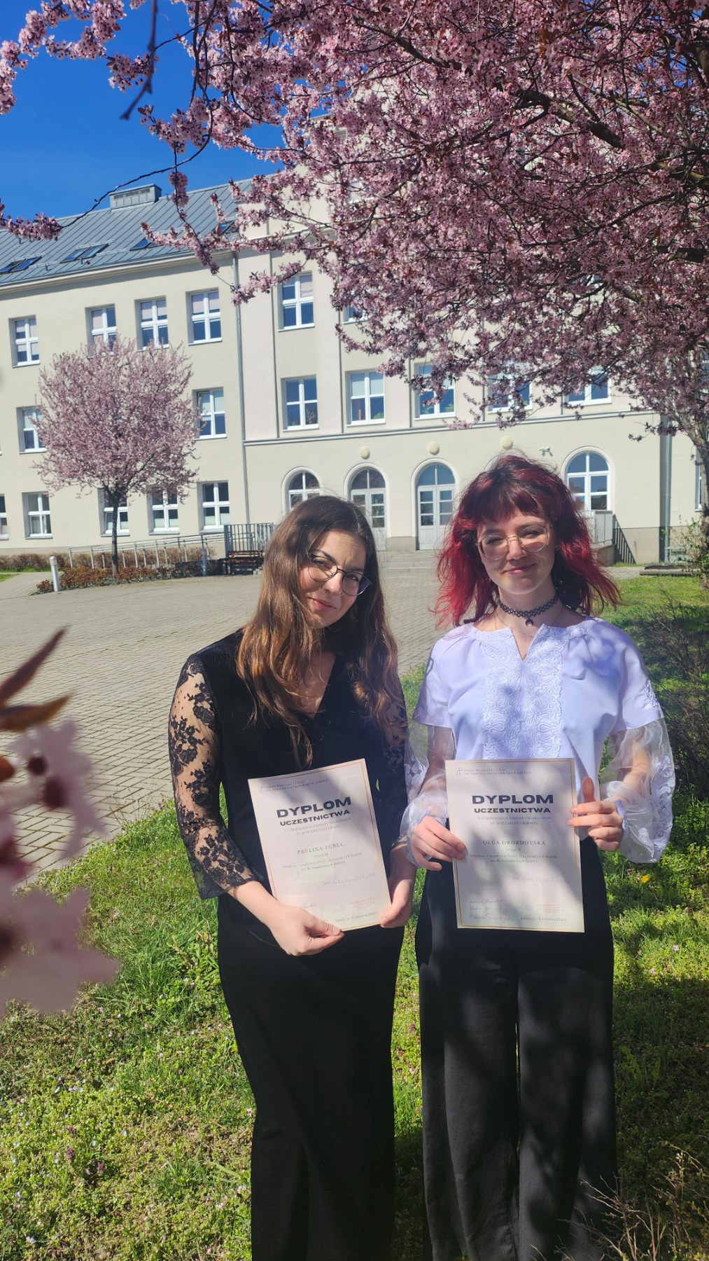 Paulina Zubel i Olga Drozdovska z dyplomami z Festiwalu Pianistycznego im. Lidii Grychtółówny. W tle budynek szkoły i kwitnące drzewa