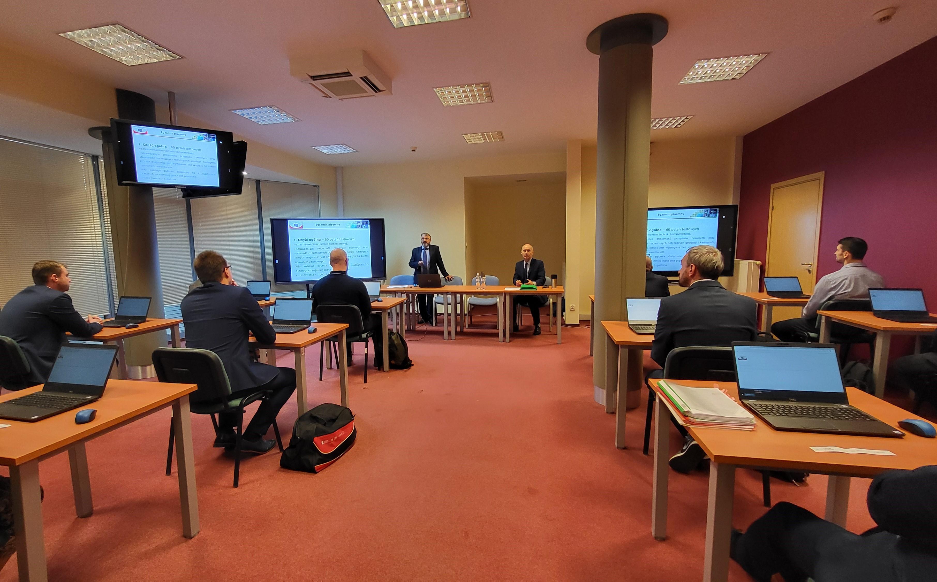 Zdjęcie przedstawia salę egzaminacyjną z członkami Komisji egzaminacyjnej i osobami przystępującymi do egzaminu siedzącymi pojedynczo przed laptopami.