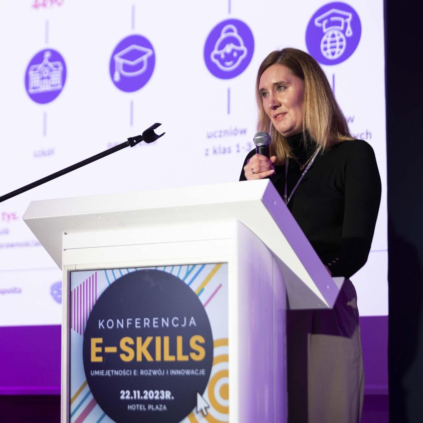 Przy mównicy stoi Pani Agnieszka Krauzowicz. W ręku trzyma mikrofon patrząc na siedzących przed nią uczestników konferencji.