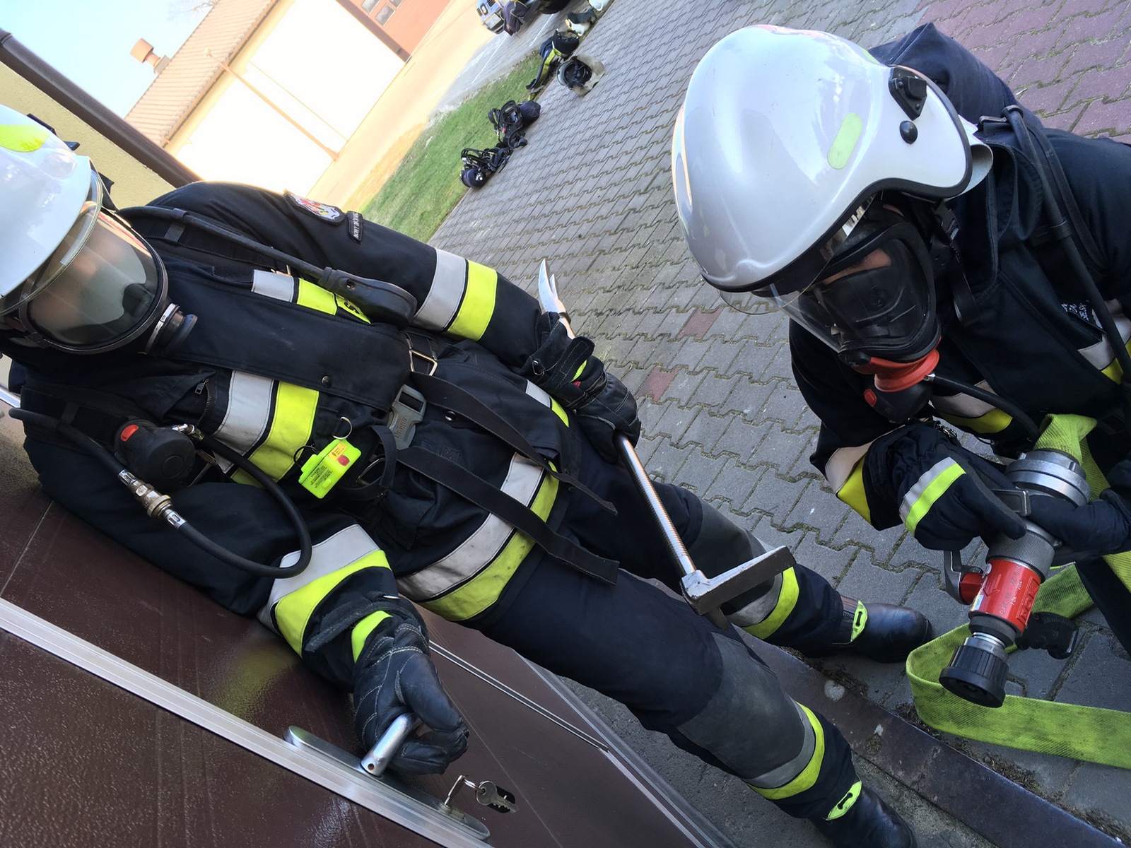 Zdjęcie przedstawia dwóch strażaków w ubraniach ochronnych, ze sprzętem gaśniczym wchodzących do budynku.