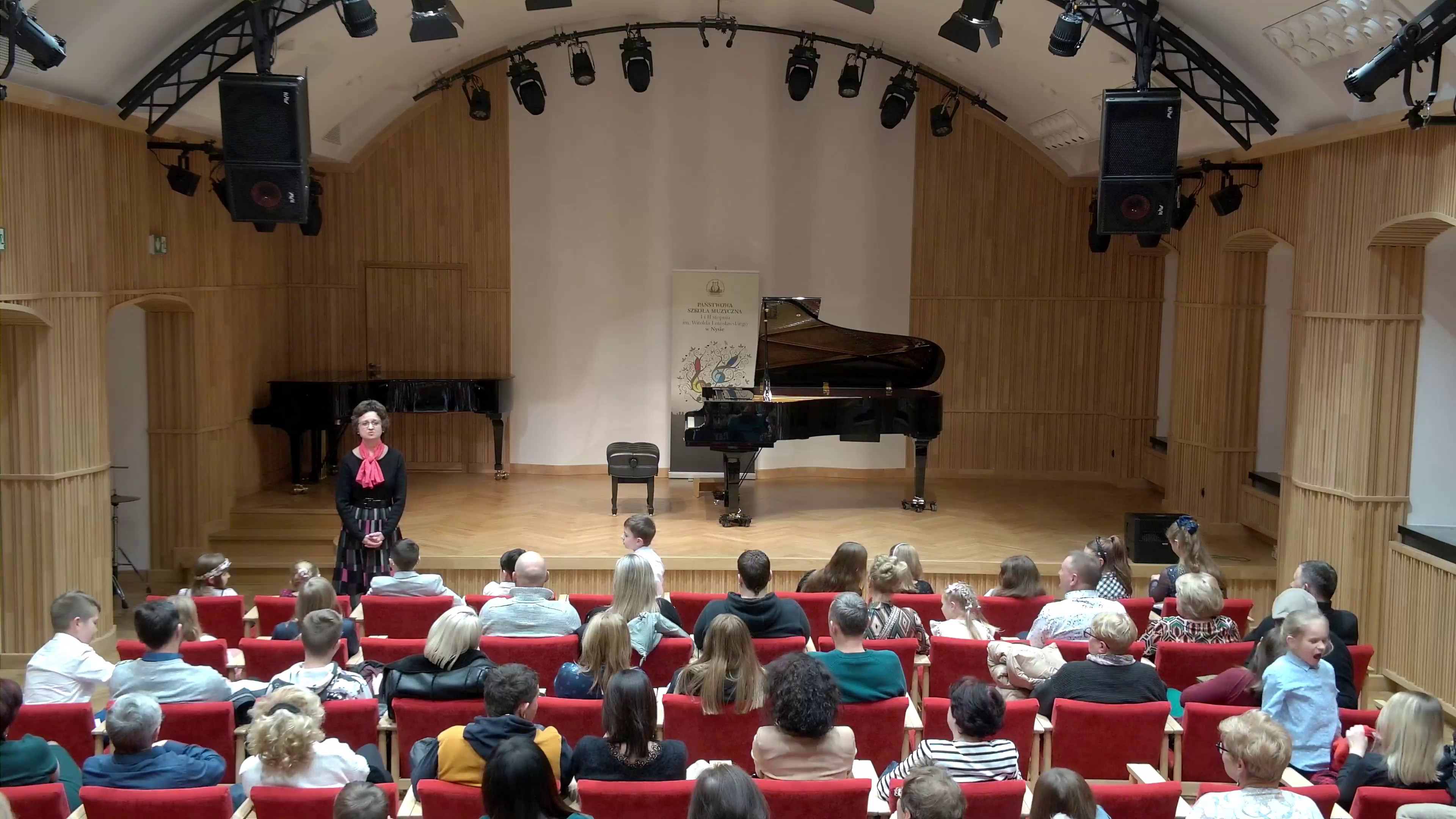 Zdjęcie. Sala Koncertowa PSM I i II stopnia w Nysie. Na pierwszym planie cztery rzędy foteli oraz siedzący w nich słuchacze. Cykl zdjęć z koncertu uczniów z klas fortepianu PSM I stopnia w Nysie - 15 grudnia 2022 - Sala Koncertowa im. Jerzego Kozarzewskiego. Występują najmłodsi pianiści. Jest to drugi koncert, który prowadzi pani Bogusława Kos. Cykl zdjęć z koncertu uczniów z klas fortepianu PSM I stopnia w Nysie - 15 grudnia 2022 - Sala Koncertowa PSM I i II stopnia w Nysie. Sala nosi imię Jerzego Kozarzewskiego. W różnych ujęciach widać młodych pianistów grających na dużym czarnym fortepianie. Na tle białej części tyłu estrady stoi baner PSM w Nysie. W rogu drugi czarny fortepian, który nie był wykorzystywany podczas koncertu.