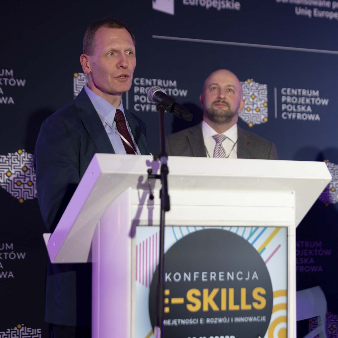 Przy mównicy stoi doktor inżynier Maciej Sajkowski. Wzrok kieruje w stronę uczetników konferencji. Mówi do mikrofonu. W tle stoi Pan Hubert Gęsiarz.