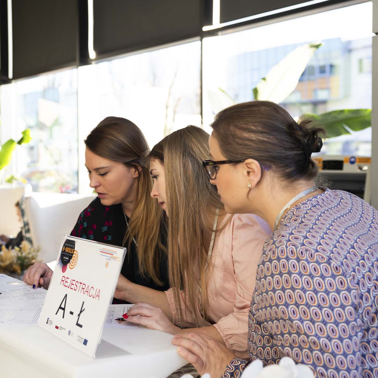 Trzy kobiety stoją przy rejestracji, wzrok kierują na listę uczestników.
