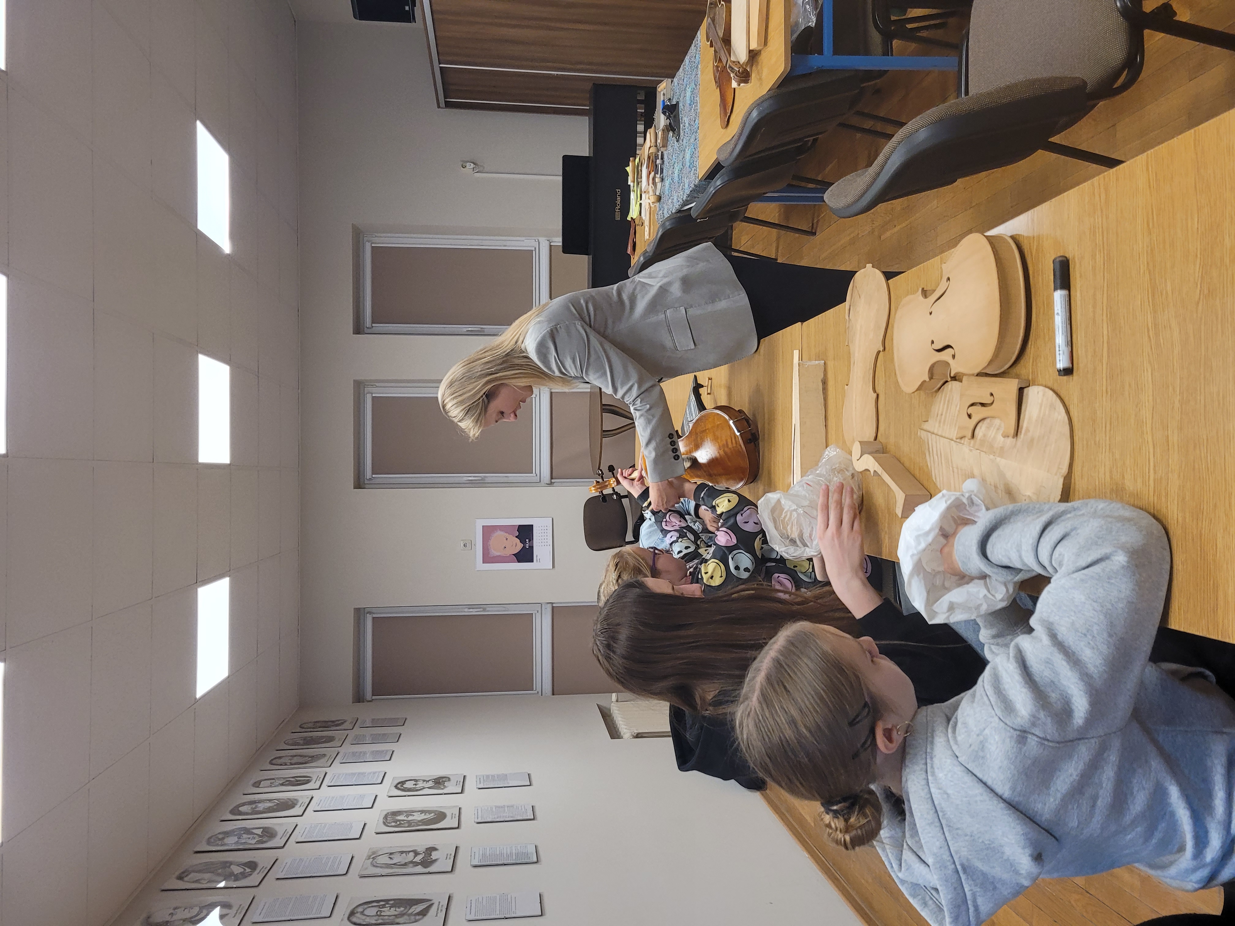 Uczniowie siedzą w ławkach i oglądają z zaciekawieniem różne elementy budowy skrzypiec. Jedna z uczennic wykonuje ruchy potrzebne przy lakierowaniu instrumentu pod okiem pani lutnik. 