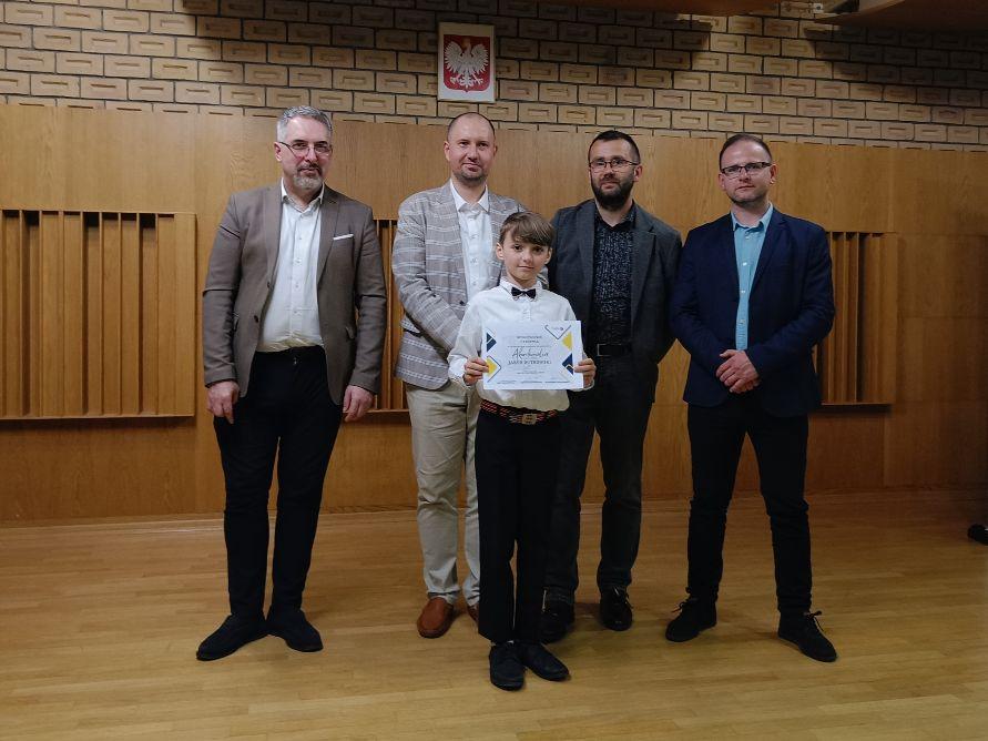 Z przodu uczeń trzymający dyplom laureata, za nim czterech członków Jury. W tle, po środku widoczne godło Polski.