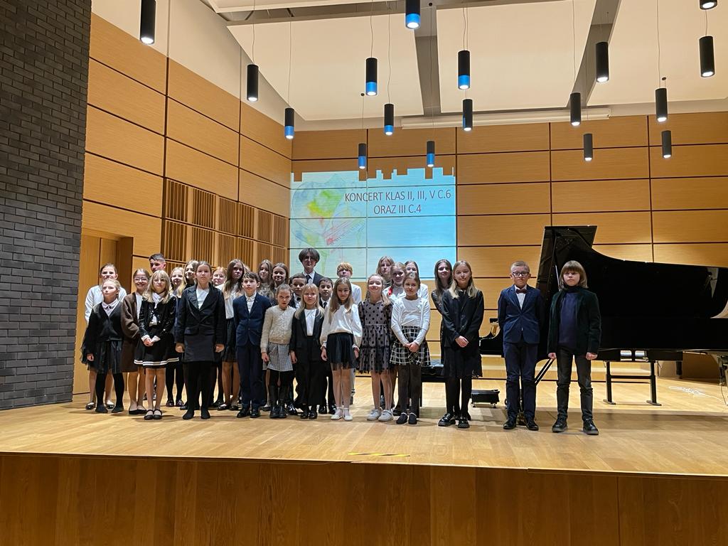 Uczniowie wraz z nauczycielami stojący w trzech rzędach na scenie sali koncertowej. Na górze widoczne wiszące lampy. Z tyłu wyświetlany na projektorze napis: "Koncert klas II, III, V c.6 oraz III c.4".