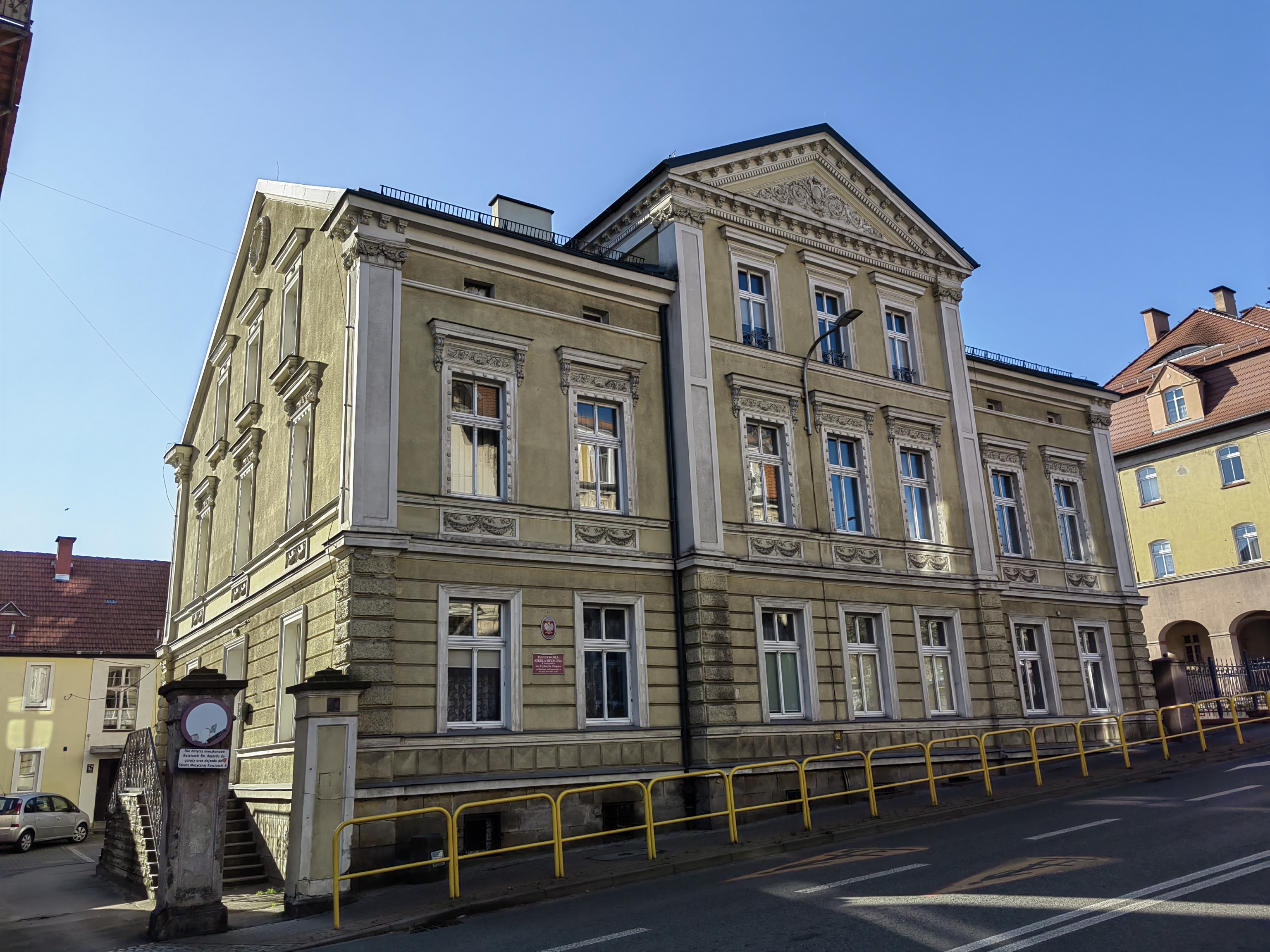 Zdjęcie wykonane w słoneczny dzień od strony ulicy, przedstawia budynek PSM I st. im. Fryderyka Chopina w Kłodzku, widoczny jest przód budynku wraz z wjazdem, oraz część ulicy, przy której znajduje się szkoła.
