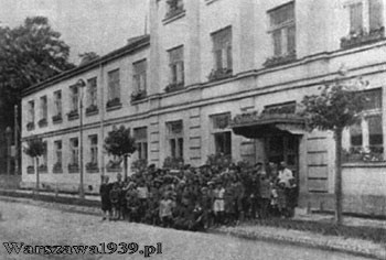 zdjęcie archiwalne, czarno-białe - piętrowy budynek szkolny w jasnym kolorze, stojący przy ulicy; przed budynkiem gromada ludzi