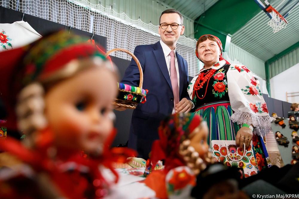 Na pierwszym planie lalka łowicka, w tle premier Mateusz Morawiecki rozmawia z jedną z gospodyń.