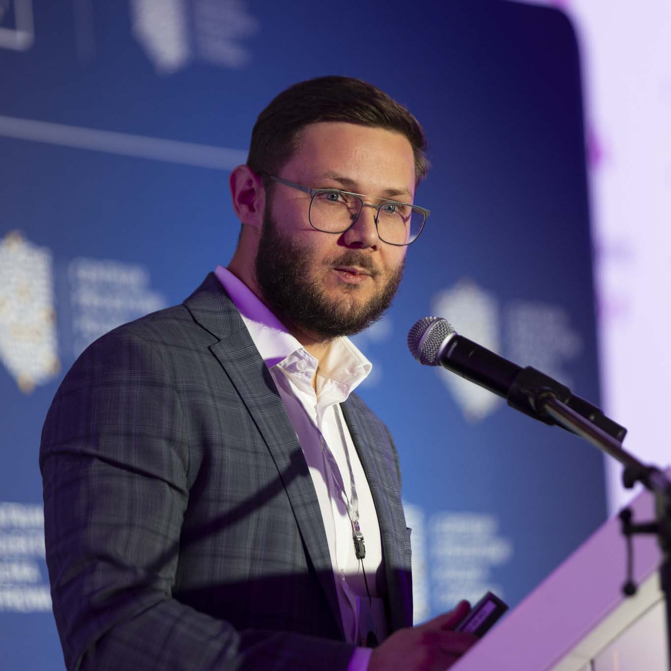 Przy mównicy stoi Pan Dawid Hoinkis. W ręku trzyma mikrofon patrząc na siedzących przed nim uczestników konferencji.