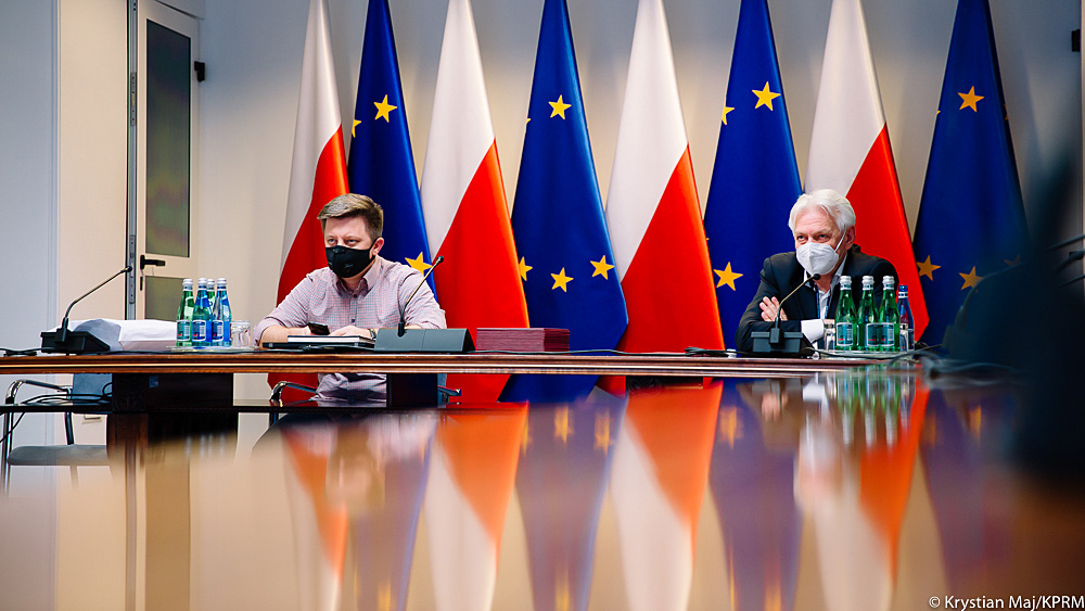 Prof. Andrzej Horban i minister Michał Dworczyk siedzą przy stole konferencyjnym podczas zdalnego posiedzenia Rady Medycznej. Obaj mają maseczki. Za nimi widać flagi Polski i Unii Europejskiej.