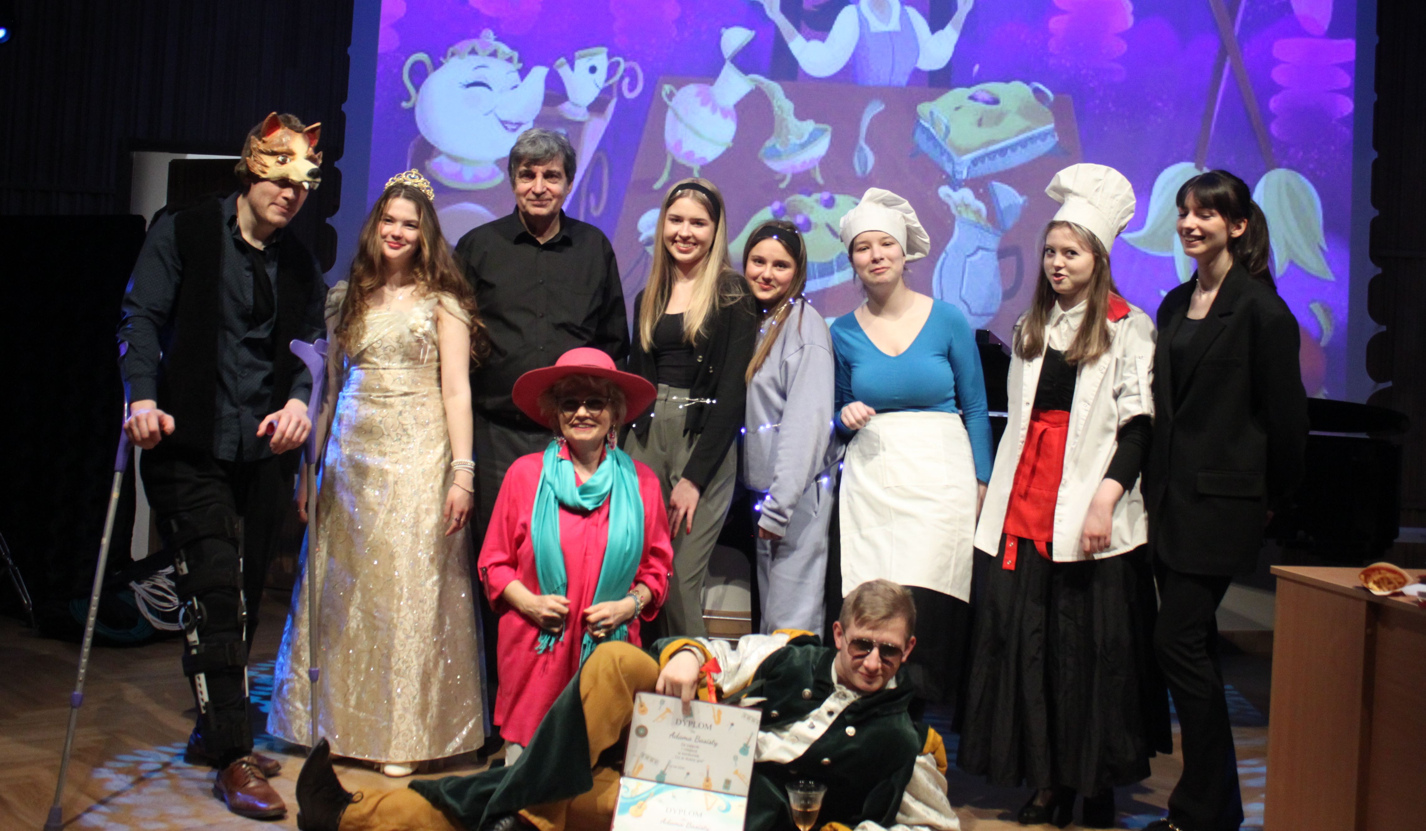 Grafika - zdjęcie. Grupa aktorów po spektaklu na estradzie pozuje do wspólnego zdjęcia. W tle fragment scenografii wyświetlony na ekranie.