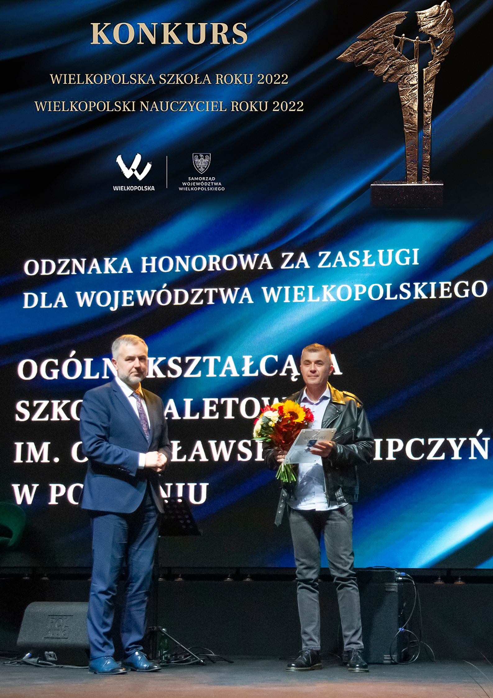 Scena, moment wręczenia odznaczenia, niebiesko czarne tło, biały napis odznaka honorowa za zasługi dla województwa wielkopolskiego, u góry w lewej części napis konkurs wielkopolska szkoła roku, po prawej statuetka konkursowa.