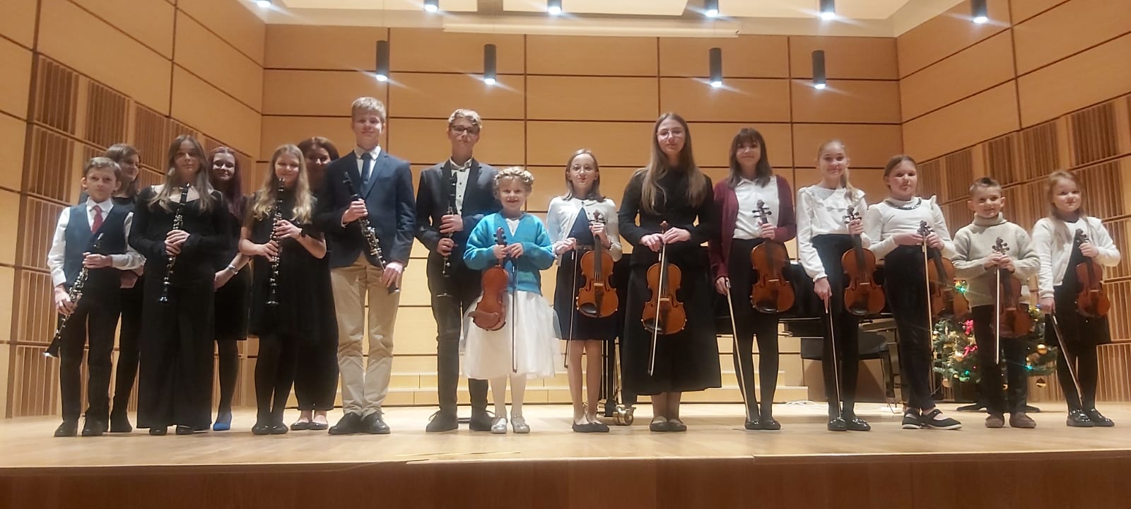 Uczniowie wraz z nauczycielami stojący w dwóch rzędach na scenie sali koncertowej. Z przodu uczniowie trzymają klarnety i skrzypce. Na górze widoczne wiszące lampy.