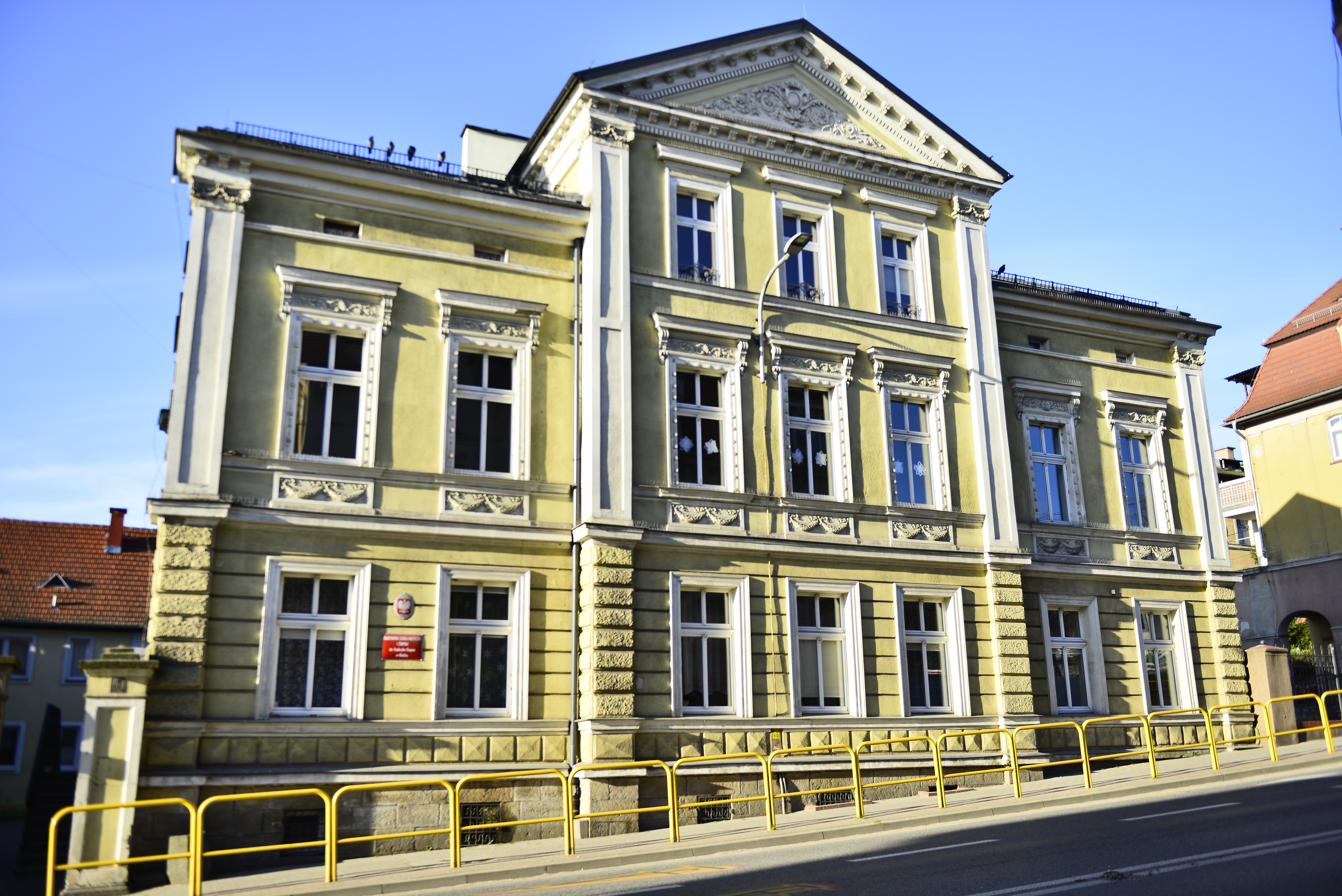 Zdjęcie wykonane w słoneczny dzień od strony ulicy, przedstawia budynek PSM I st. im. Fryderyka Chopina w Kłodzku, widoczny jest przód budynku wraz z wjazdem, oraz część ulicy, przy której znajduje się szkoła.