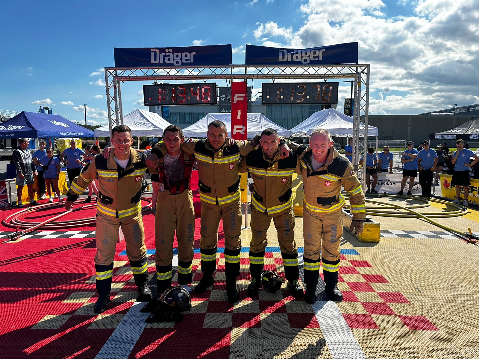 5 zawodników w mundurach strażackich trzyma się za zramiona, w tle tablica sportowa z wysiwetlonymi wynikami 1:13.48 i 1.13.78
