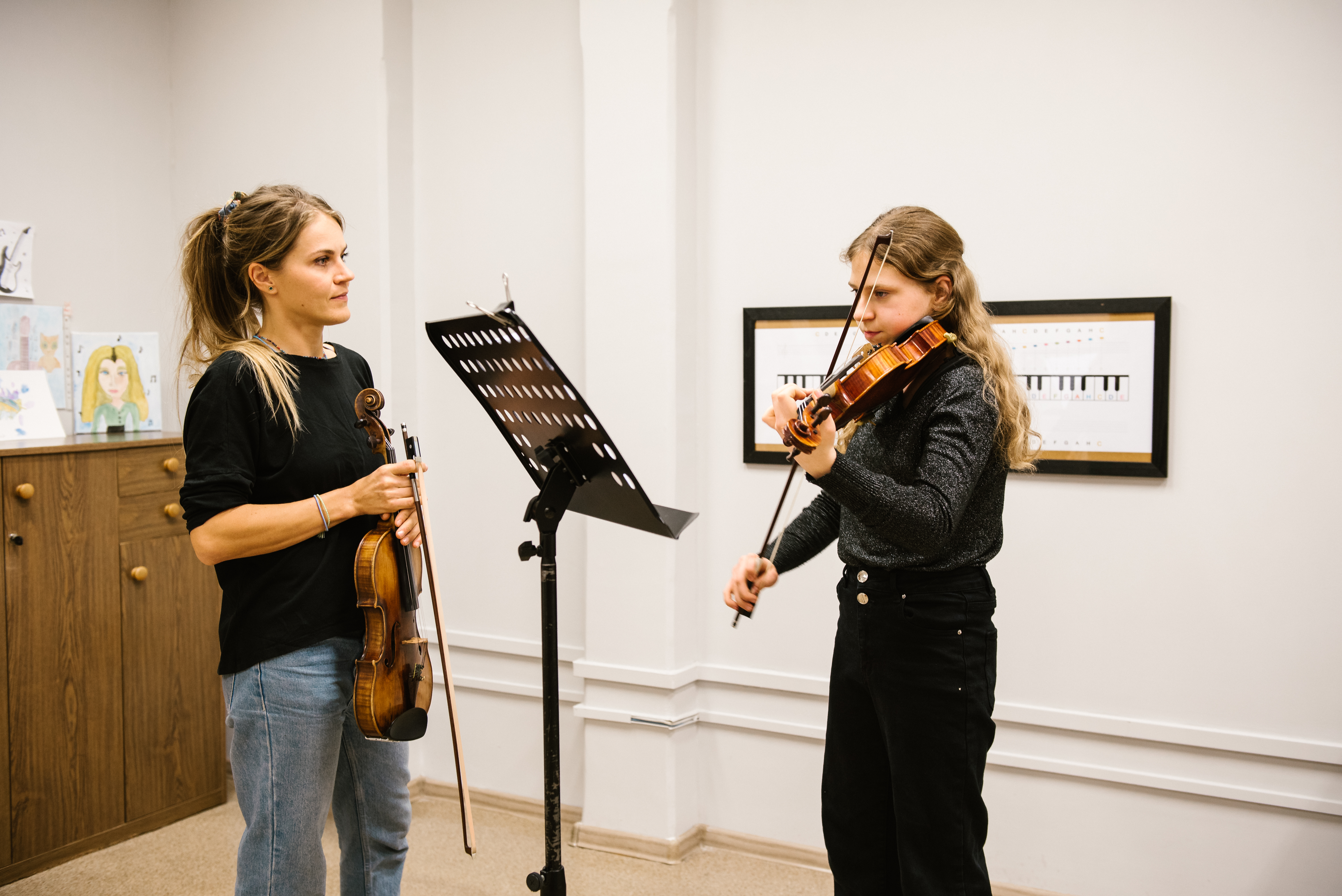 Na zdjęciu znajduje się pani Edyta Kornafel oraz uczennica Lena Baryła. Nauczycielka trzyma w rękach skrzypce i uważnie słucha gry uczennicy na skrzypcach z nut znajdujących się na czarnym pulpicie. W tle stoi brązowa komoda z rysunkami, na ścianie wisi meloplast w czarnej ramce. 