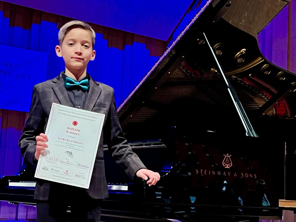 Na zdjęciu widoczny jest elegancko obrany uczeń, który w prawej ręce trzyma dyplom konkursowy. Lewą dłoń opiera o stojący obok fortepian.