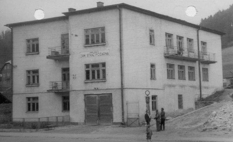 Widok poprzedniego budynku głównego strażnicy JRG Krynica. Bryła budynku w kształcie sześcianu osadzona na zboczu. Obok budynku stoi kilka osób. w tle widać las.