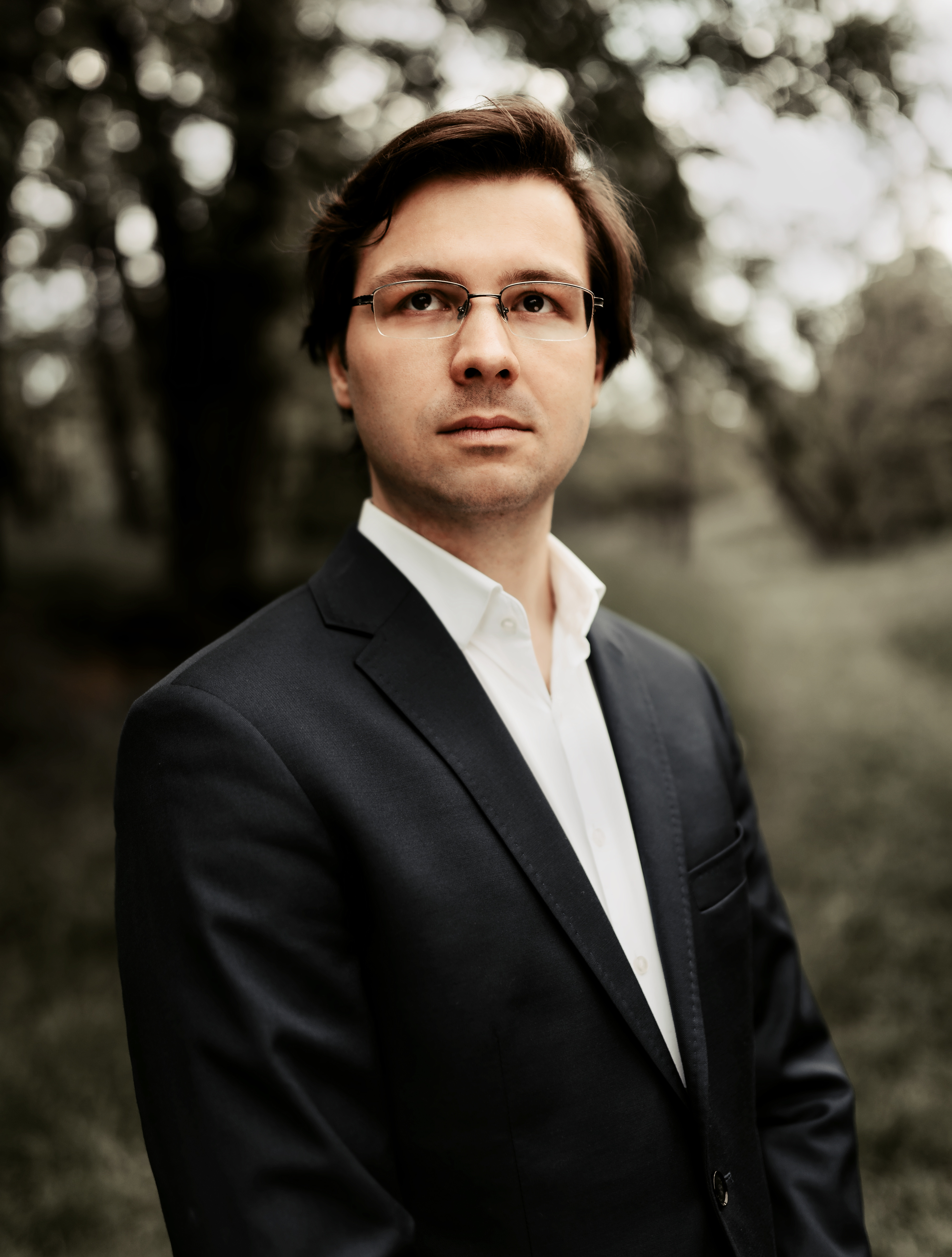  Zdjęcie pianisty Wojciecha Pyrć. Artysta w jasnej koszuli i garniturze na tle krajobrazu drzew i traw.