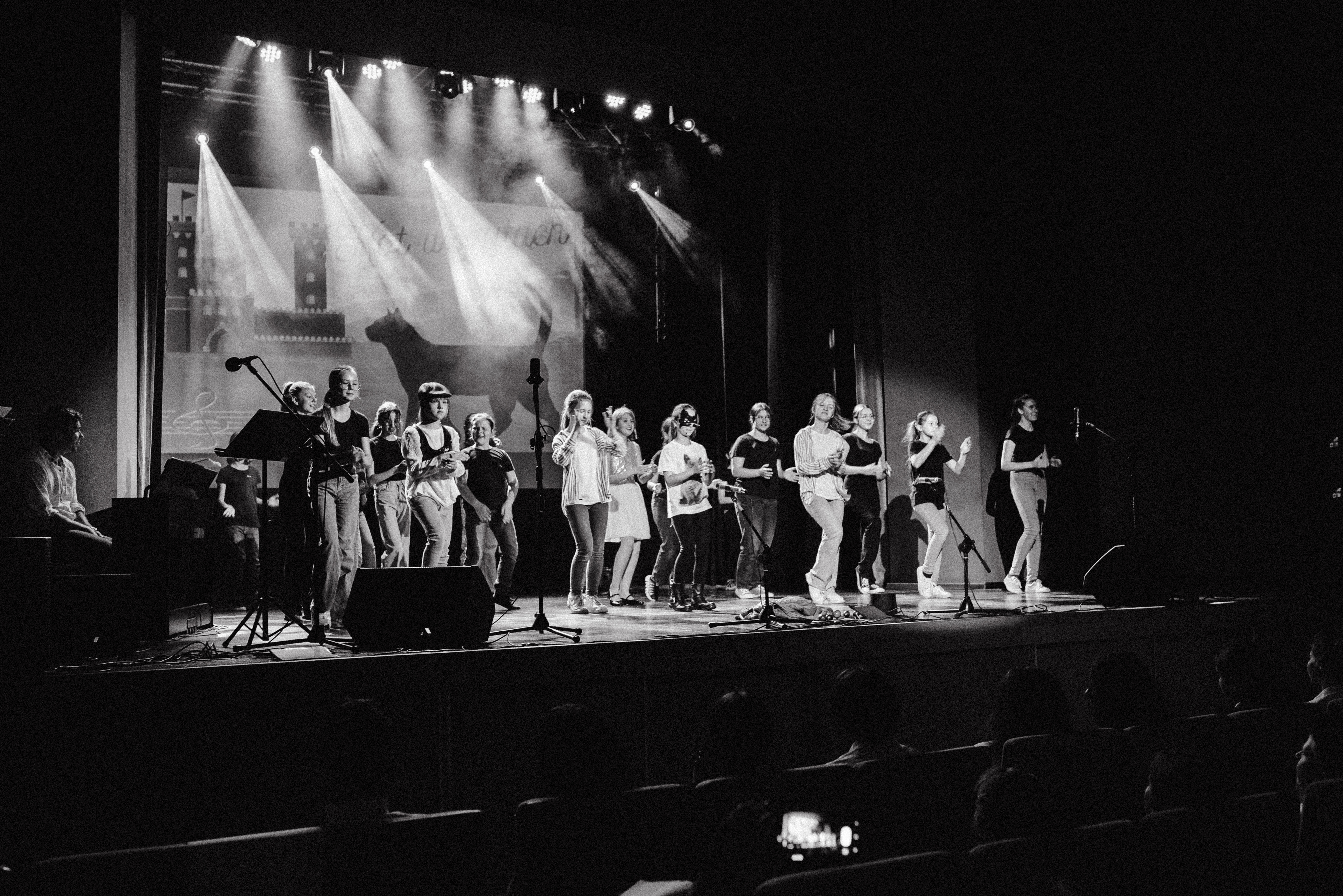 Czarno-białe zdjęcie przedstawia tańczącą grupę dzieci na scenie. Widoczna jest także siedząca publiczność. 