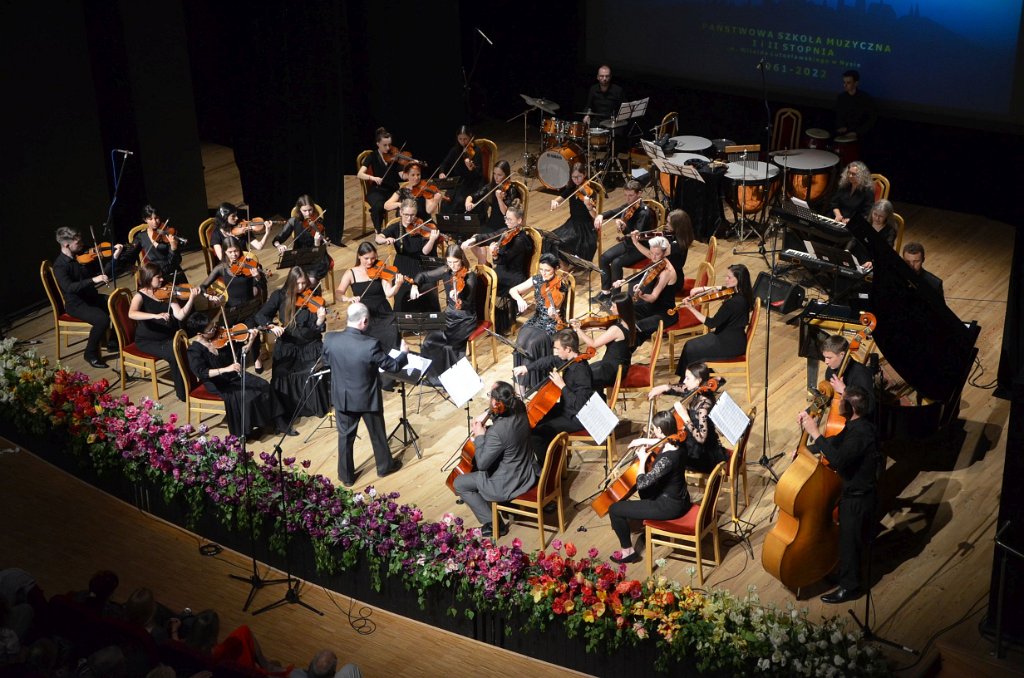 Zdjęcie: Jubileusz 60-lecia PSM w Nysie - II Koncert Jubileuszowy - 14 maja 2022 - Nyski Dom Kultury - scena - Orkiestra Kameralna pod dyrekcją Krzysztofa Sali. W skład orkiestry wchodzą uczniowie, nauczyciele i absolwenci szkoły. W tle wyświetlone na wiszącym ekranie logo jubileuszu. Na całym obrzeżu sceny bukiety różnokolorowych kwiatów. Kliknięcie na zdjęcie przeniesie do prezentacji cyklu zdjęć z koncertu.