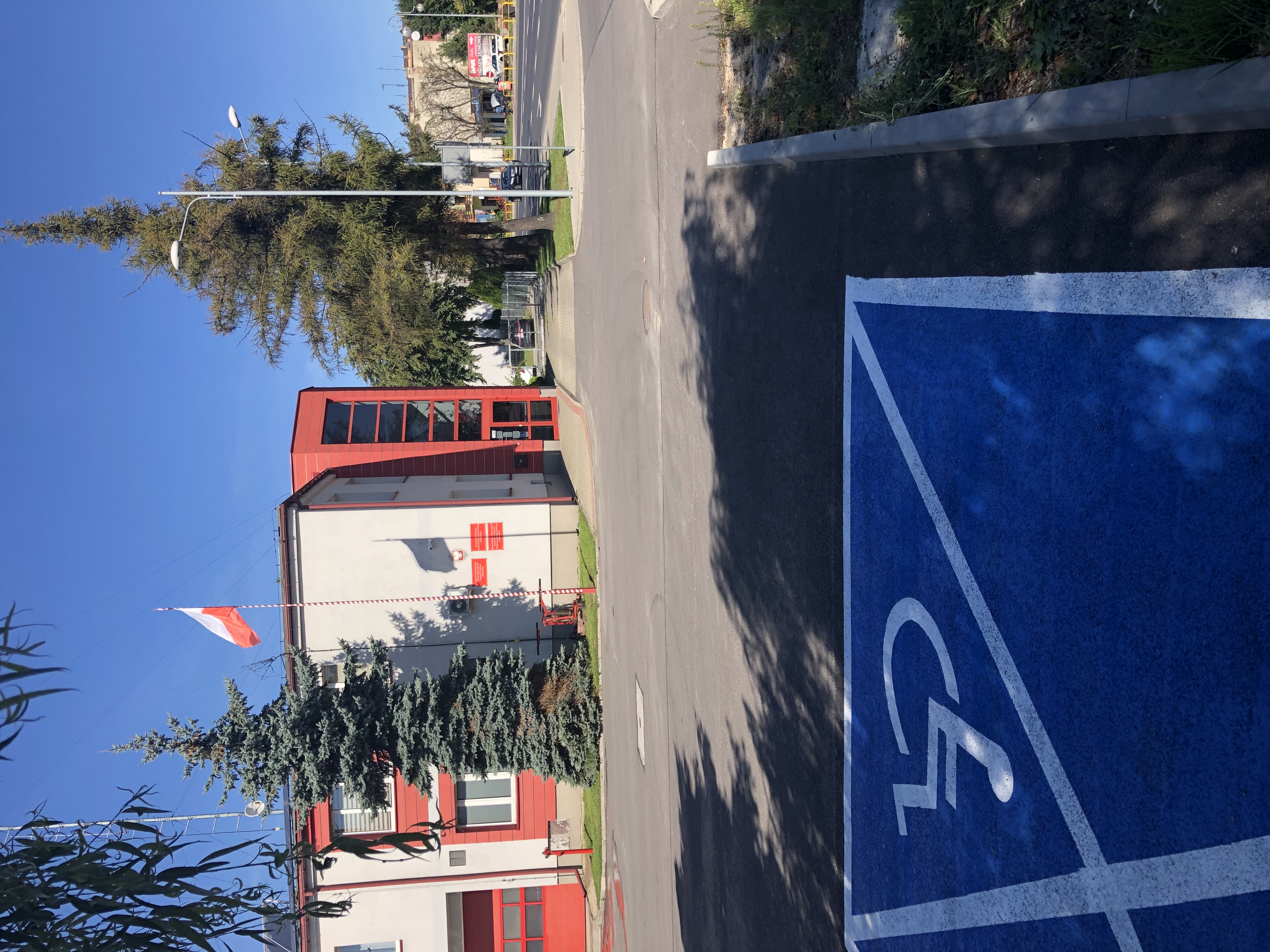 Parking z namalowanym na niebiesko miejscem postojowym dla osób z niepełnosprawnością. W tle budynek komendy szary z czerwonymi elementami.