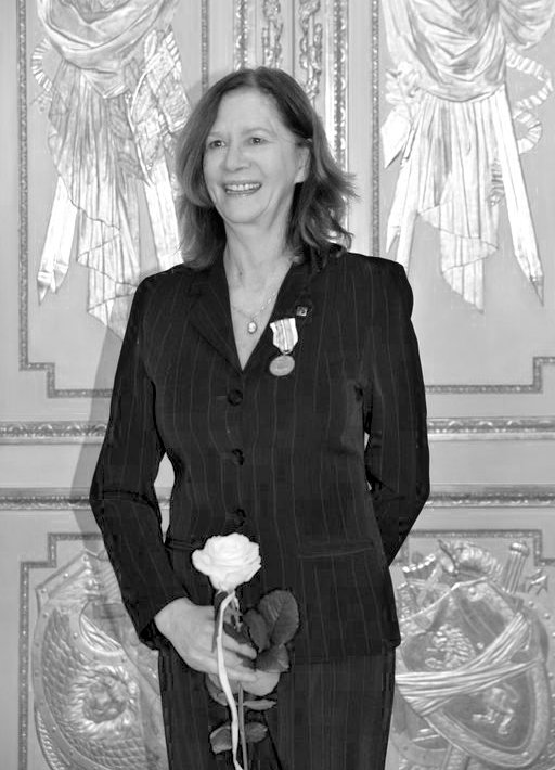 Czarno-białe zdjęcie, uśmiechnięta kobieta w eleganckim stroju, w ręku trzyma kwiat, na piersi przypięty medal