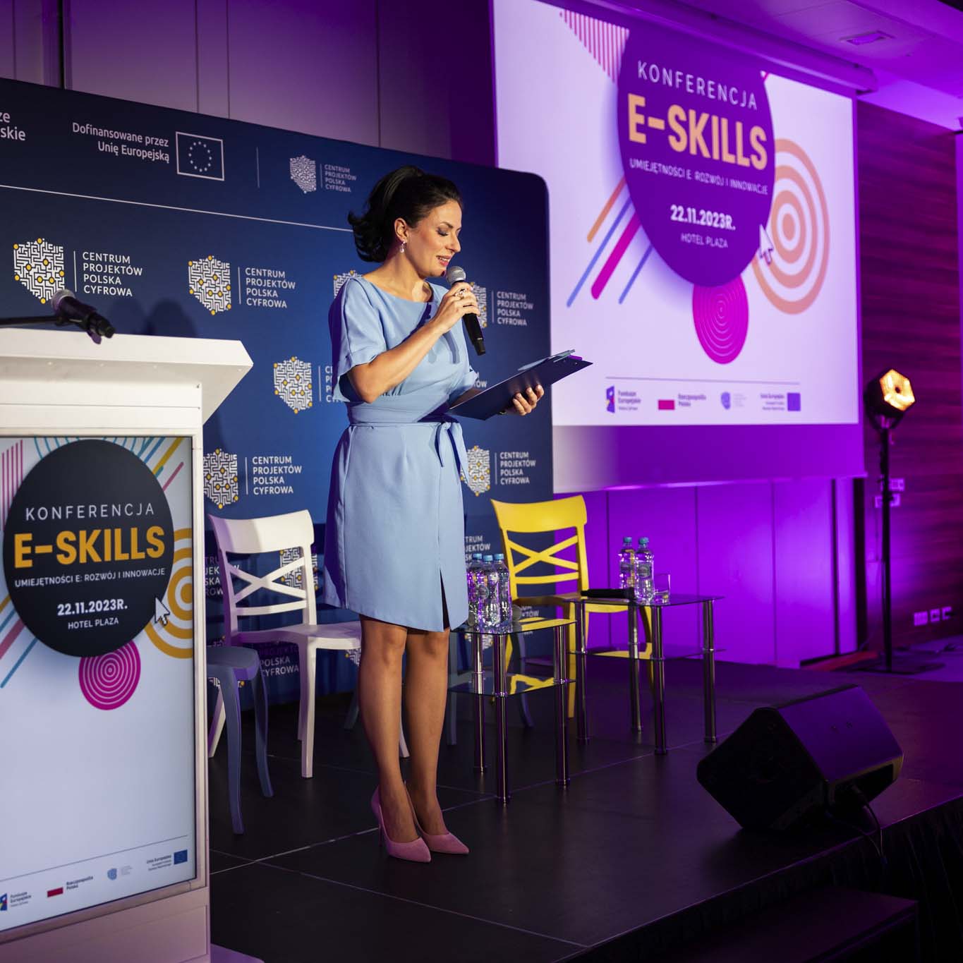 Pani Anna Butrym stoi na środku podium trzyma w ręku mikrofon. Wzrok kieruje na notatki dotyczące szczegółów konferencji.