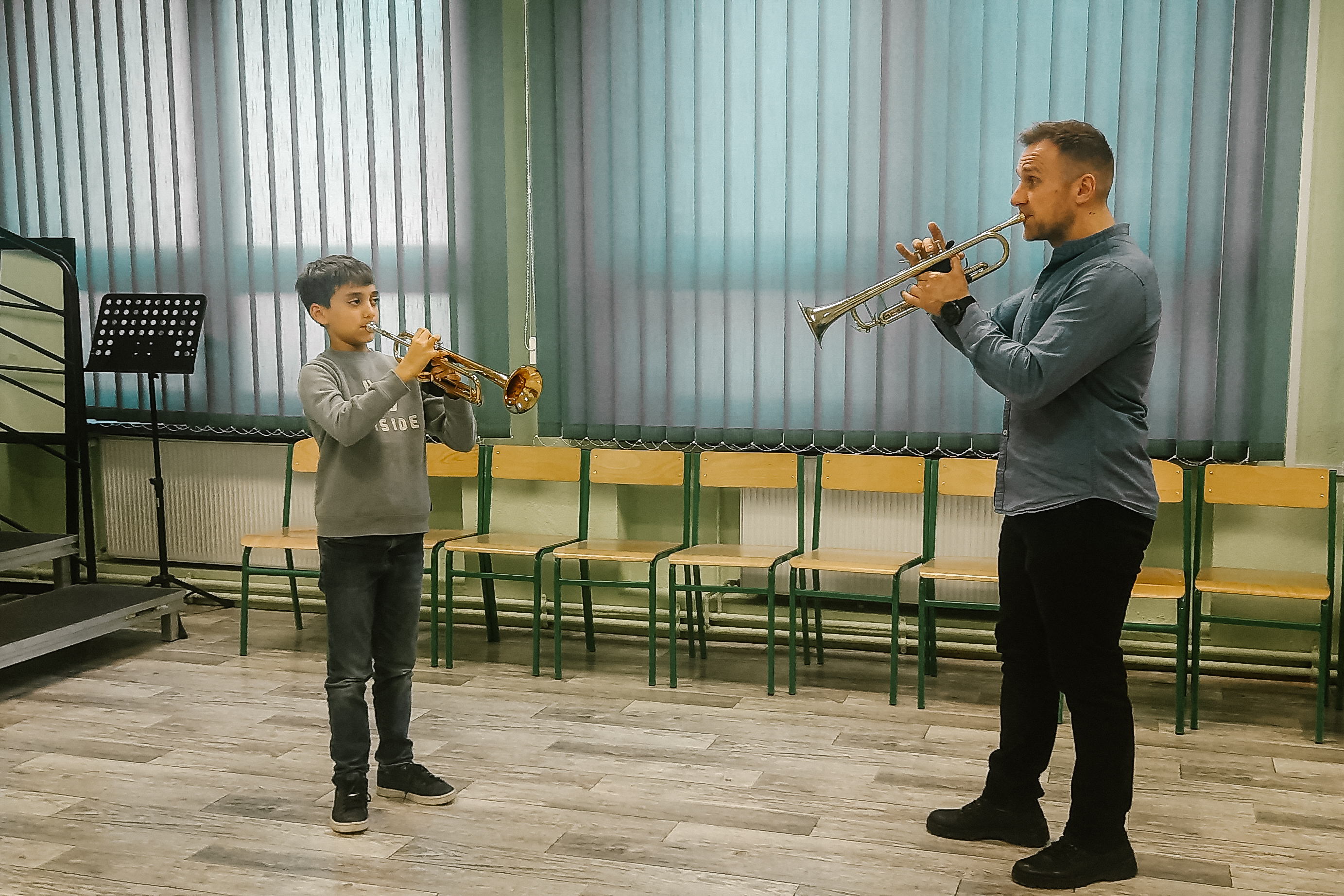 Na zdjęciu znajduje się nauczyciel Paweł Ficoń oraz uczeń Mateusz Forgiel grający na trąbkach. W tle znajdują się krzesła klasowe oraz czarny pulpit.