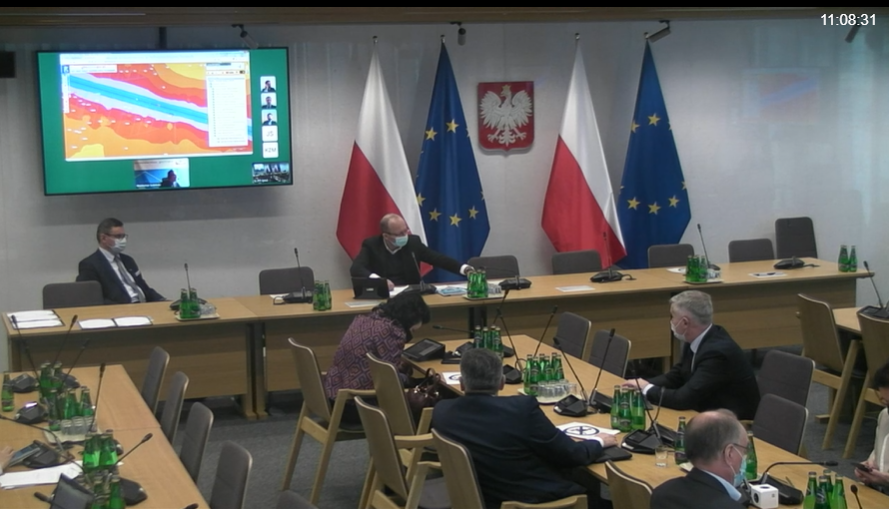 Zrzut ekranu z transmisji posiedzenia Sejmowej Komisji Infrastruktury, na którym widać posłów siedzących w sali oraz ekran pokazujący zdalną prezentację Geoportalu