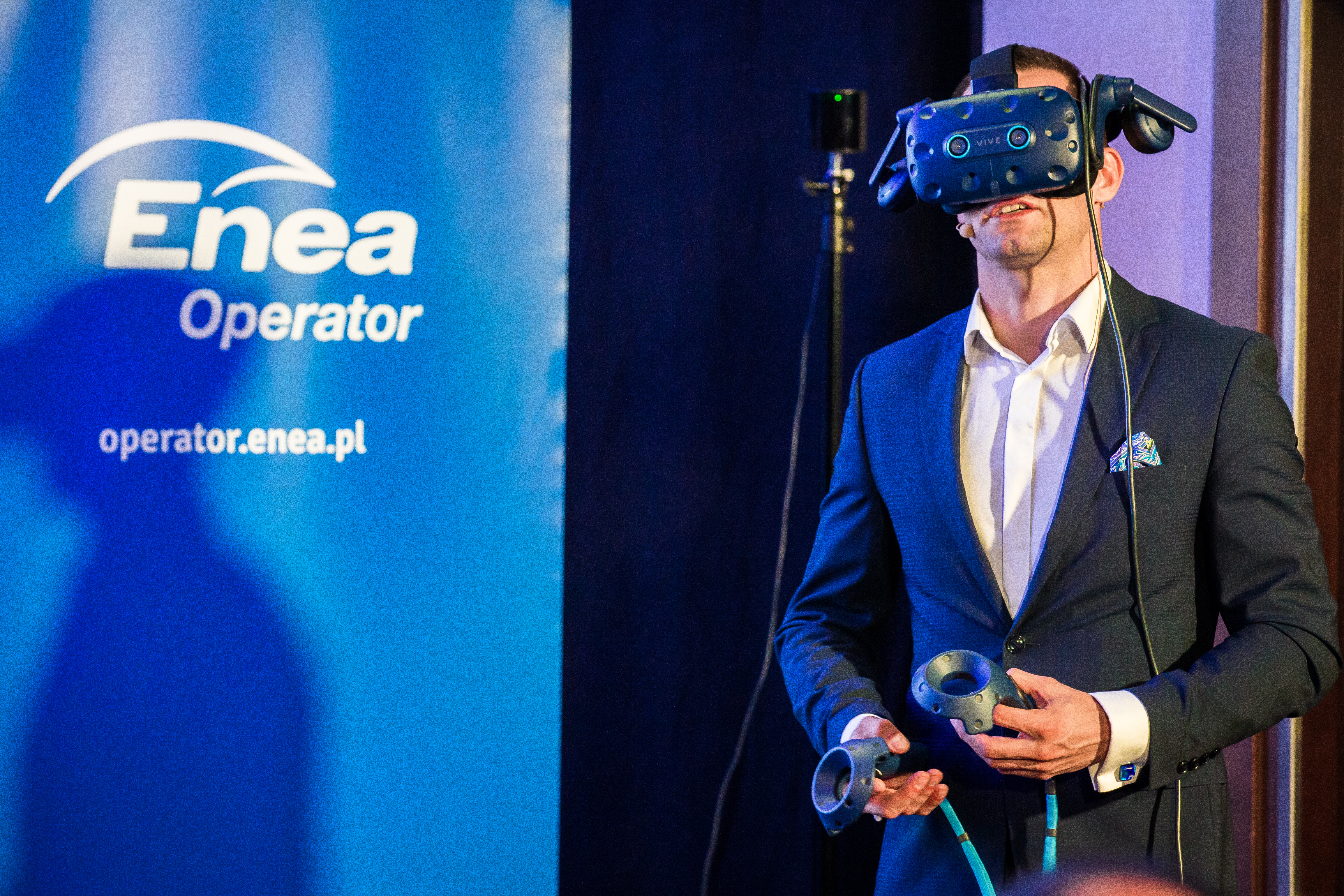 Prezenter pokazuje działanie systemu VR. Prezenter ma założone specjalne gogle i podłączone czujniki. Po lewo wyświetlone logo ENEA Operator.