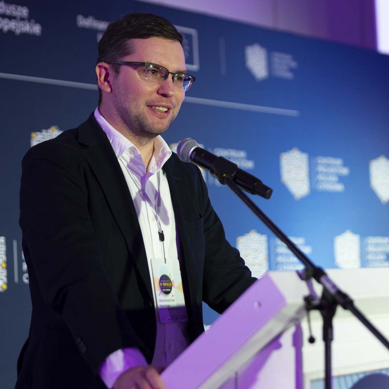 Przy mównicy stoi Pan Daniel Duda. Wzrok kieruje w stronę uczestników konferencji. Mówi do mikrofonu.