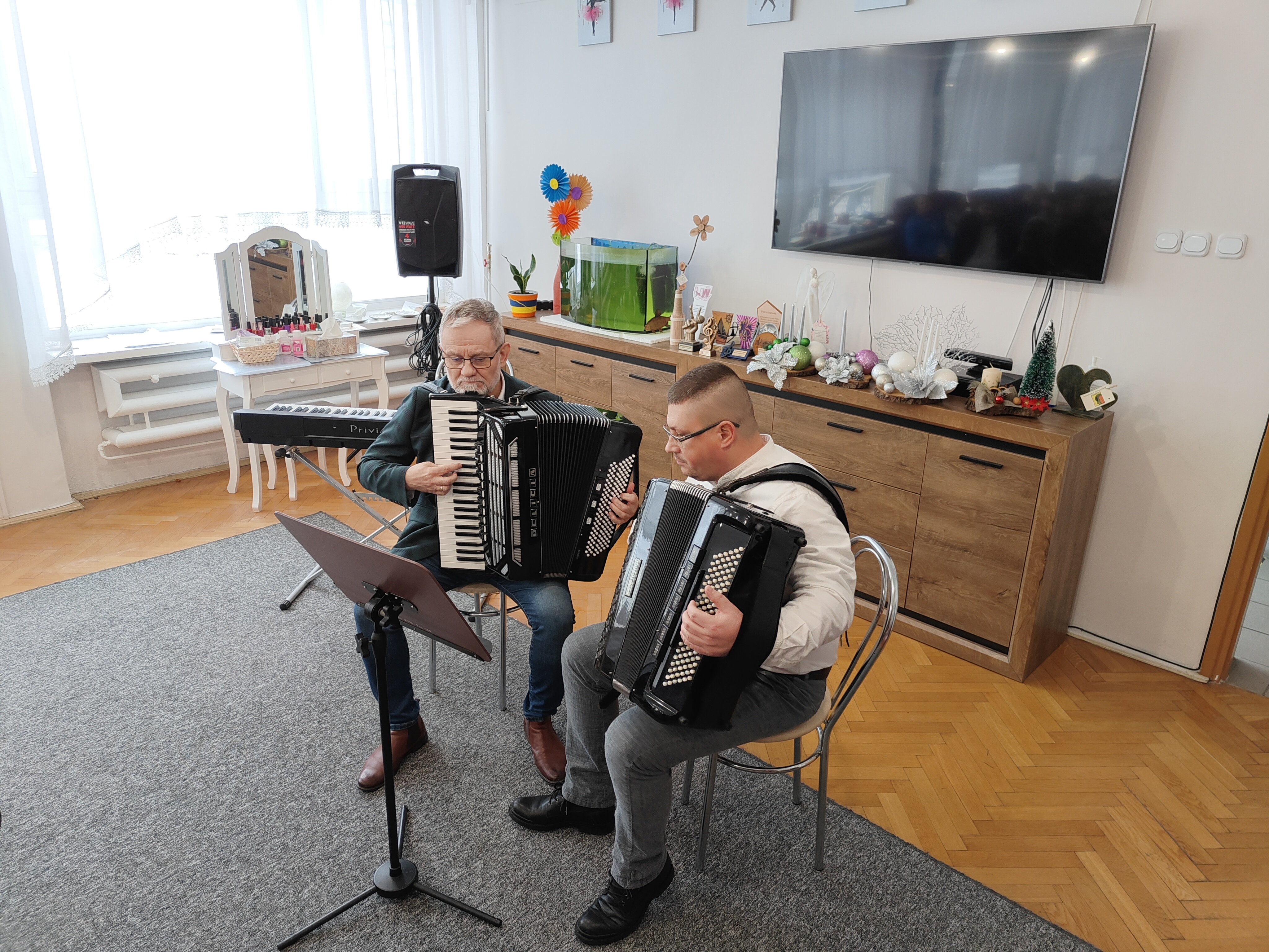 Na zdjęciu widać dwóch mężczyzn grających na czarnych akordeonach. Obaj siedzą na jasnych krzesłach. Przed nimi stoi pulpit na nuty. Mężczyzna siedzący z prawej strony jest ubrany w niebieskie spodnie oraz białą koszulę. Mężczyzna siedzący z lewej strony jest ubrany w niebieskie spodnie, białą koszulę oraz ciemną marynarkę. W tle widać brązową komodę z dekoracją świąteczną, telewizor zawieszony na jasnej ścianie, czarny instrument klawiszowy na statywie oraz kolumnę głośnika na statywie.