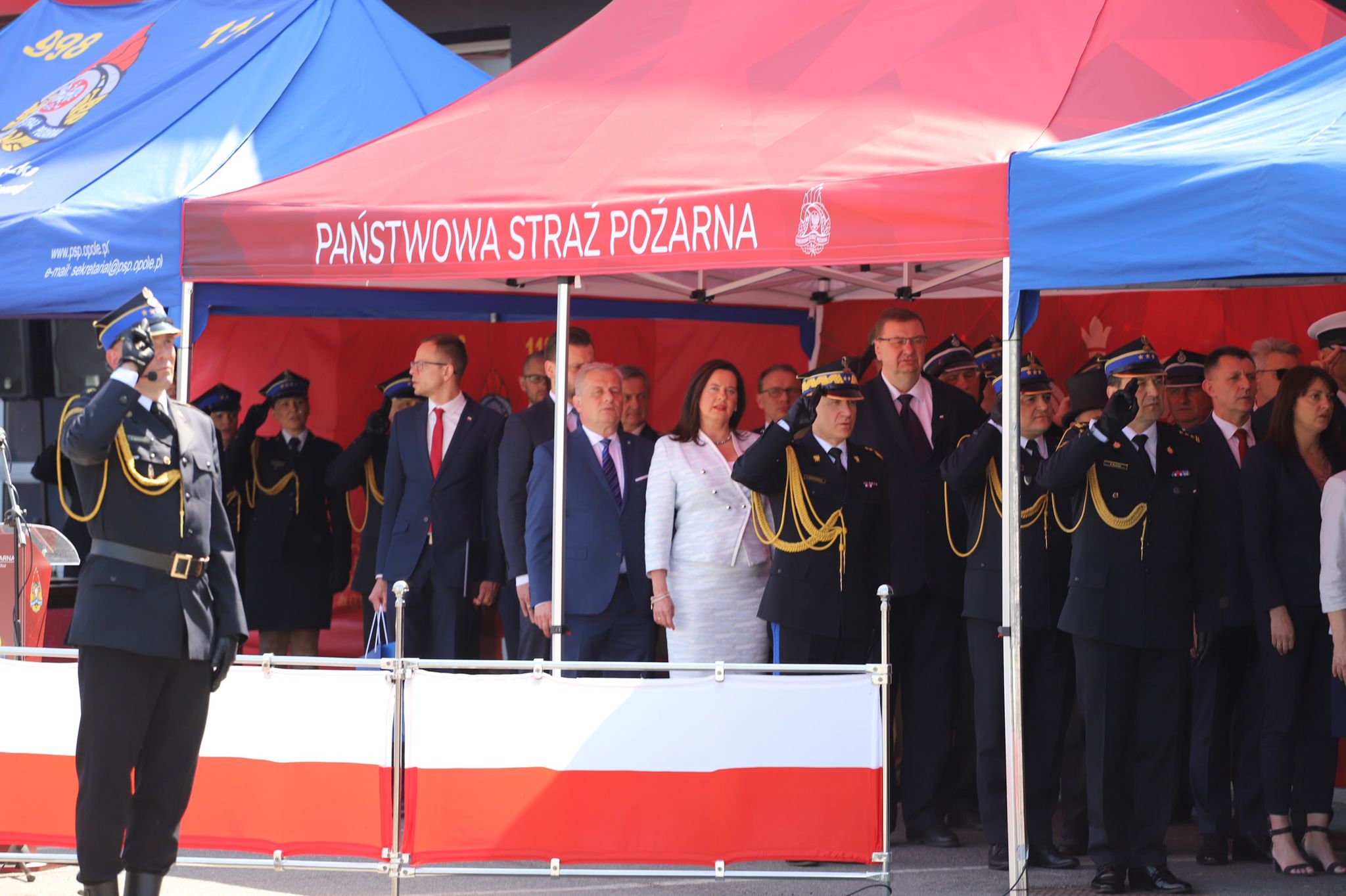 Zebrani goście pod namiotem koloru czerwonego z napisem Państwowa Straż Pożarna podczas hymnu Polski 