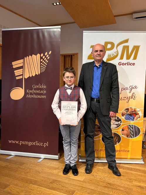 Zdjęcie przedstawia ucznia trzymającego dyplom laureata, obok niego stoi nauczyciel. W tle plakaty promujące konkurs.